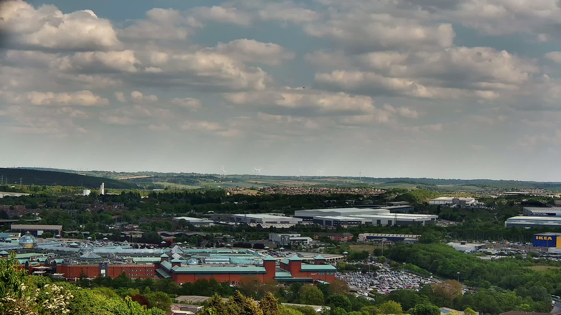 A sprawling shopping center and industrial park sit nestled amongst green hills under a partly cloudy sky.