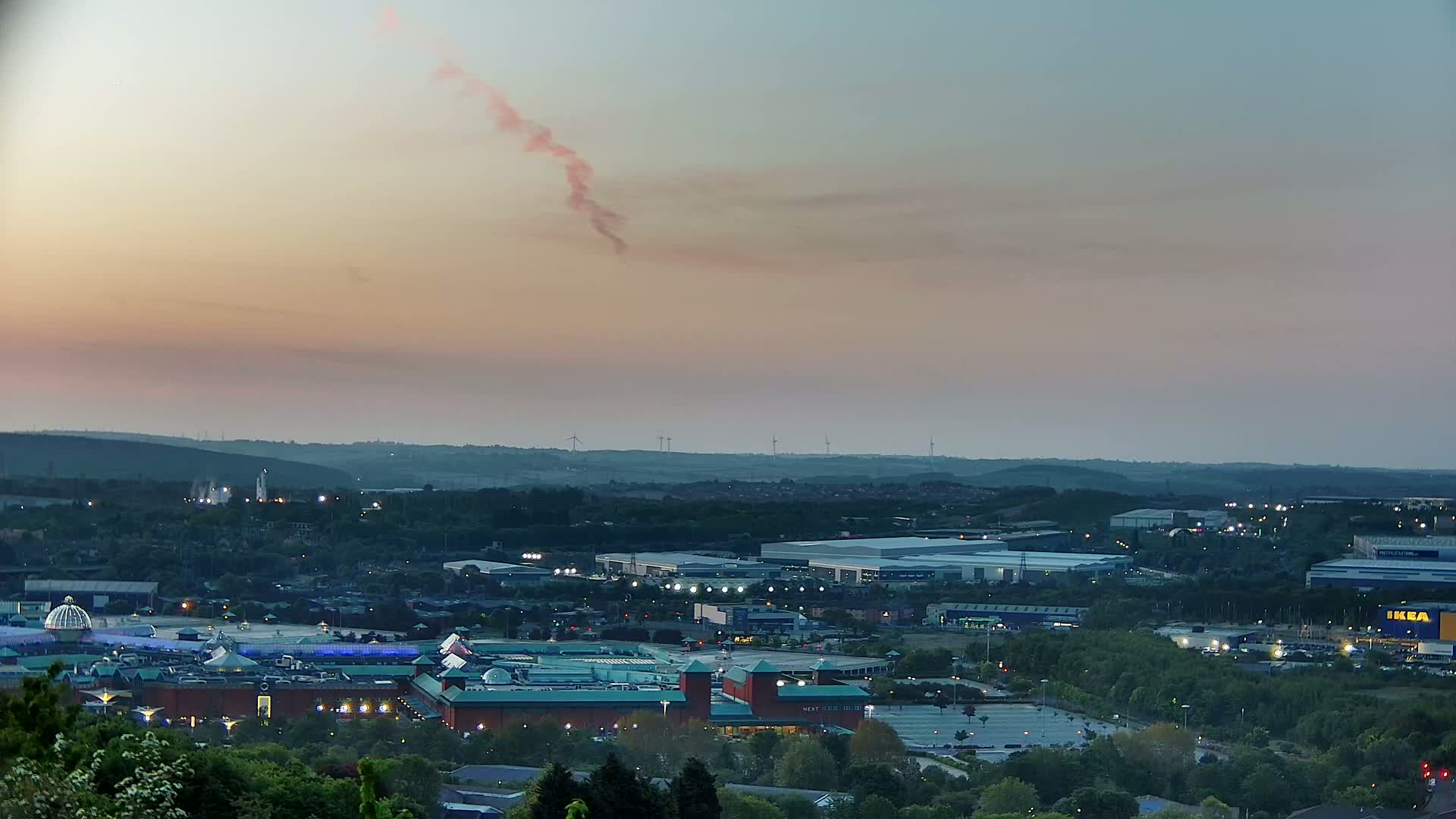 A twilight view of a large shopping center complex and industrial park, nestled among green hills and dotted with wind turbines under a pale pink and blue sky.