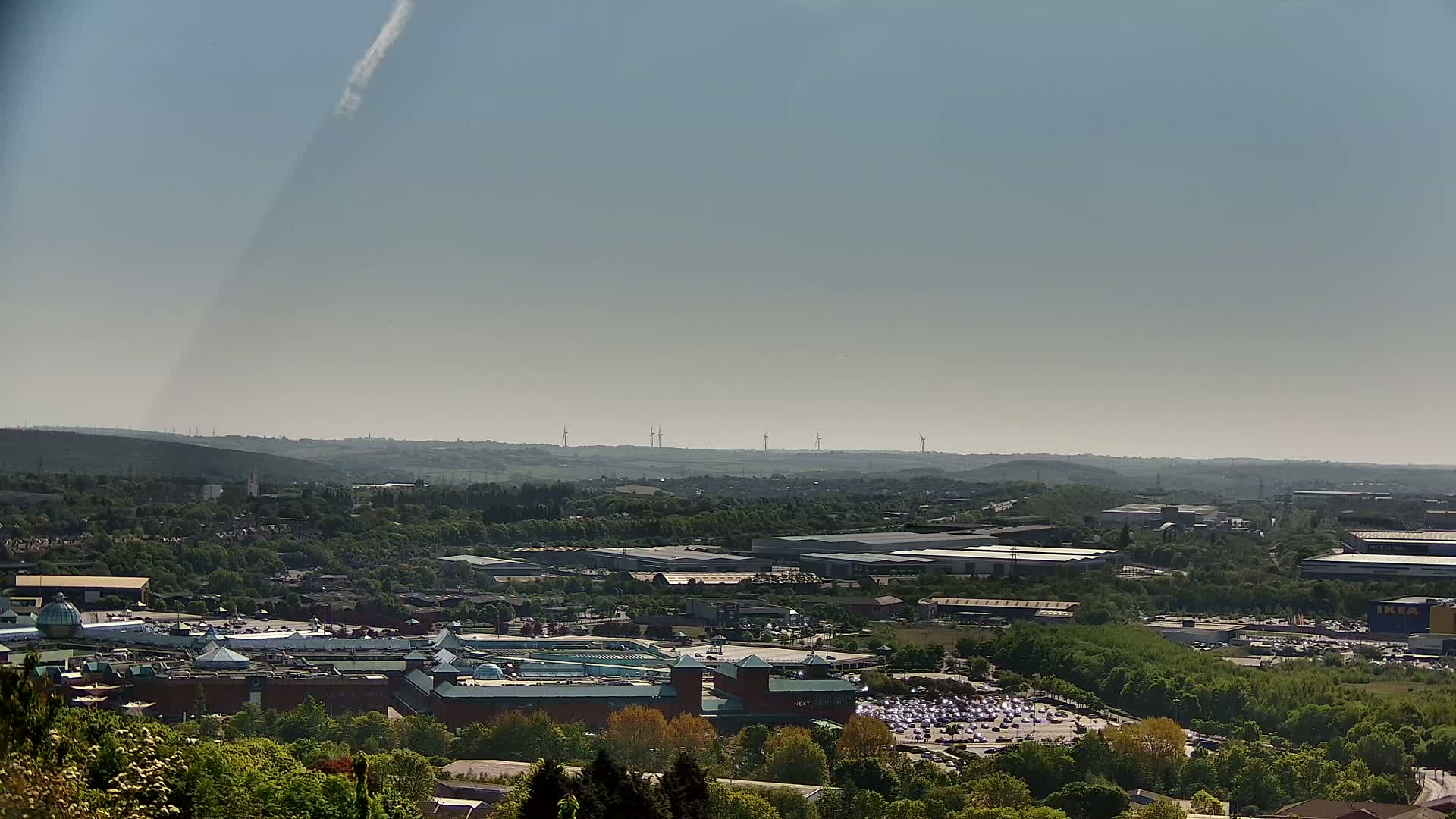 A hazy, sunny day reveals a sprawling landscape of industrial buildings, a large shopping center, and extensive parking lots, all nestled amongst green hills and trees, with wind turbines visible in the distance.