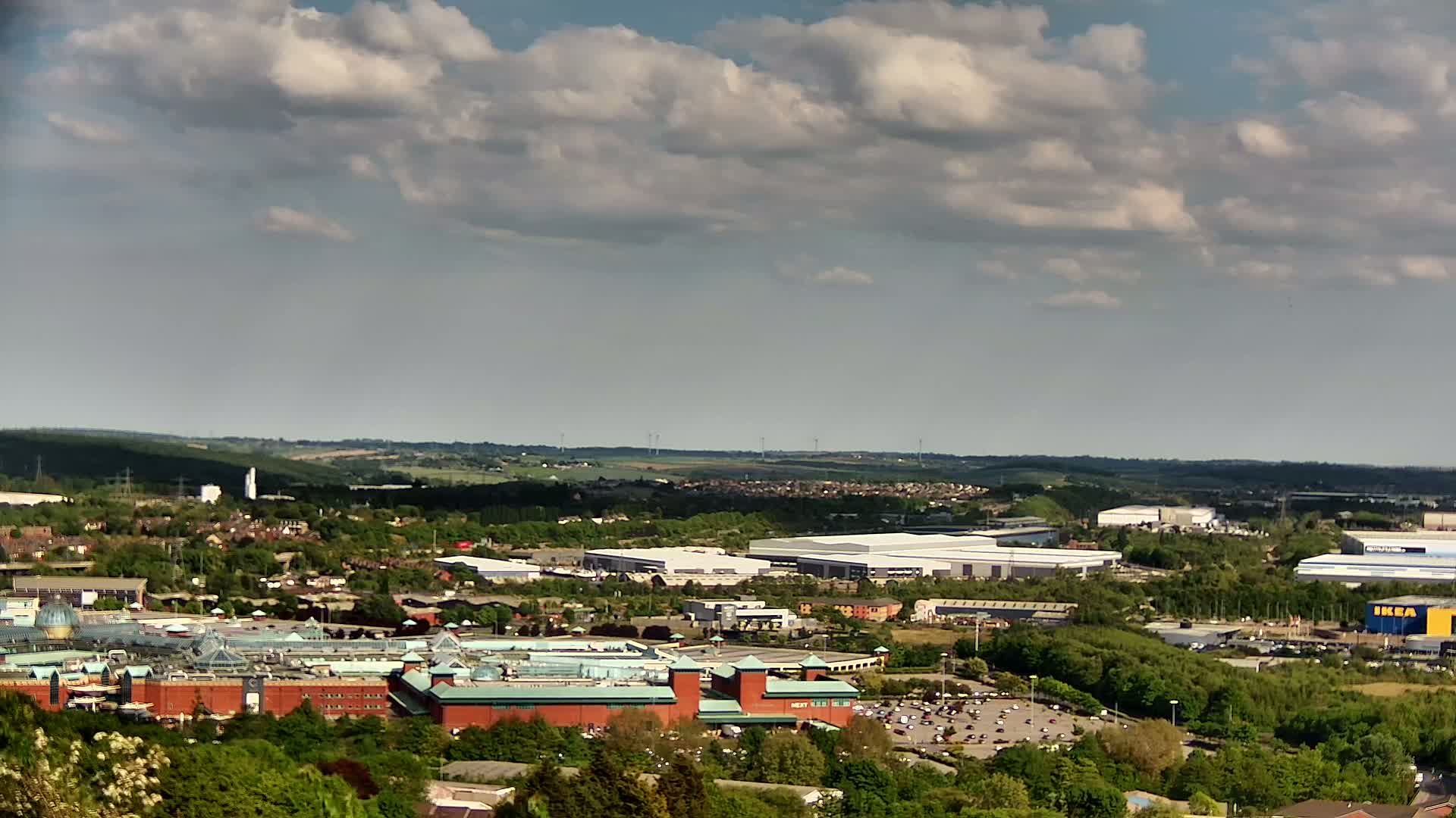A sprawling suburban landscape under a partly cloudy sky features a large shopping mall and numerous industrial buildings amidst green trees and residential areas.