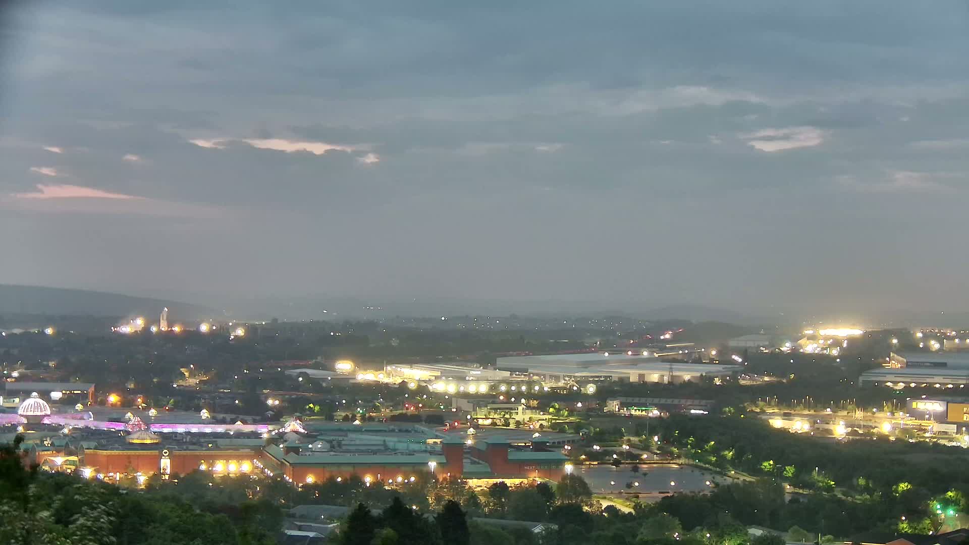 A nighttime, overcast view shows a sprawling city with illuminated buildings, parking lots, and green spaces, viewed from an elevated vantage point.