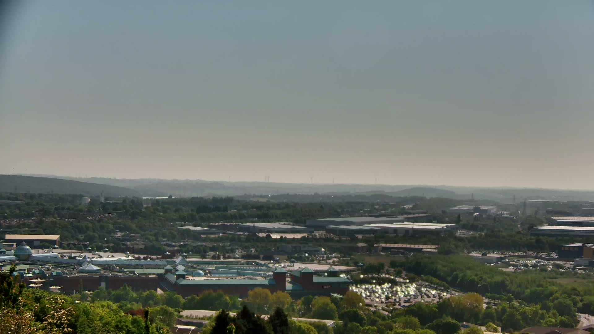 A hazy view shows a large shopping center nestled among green trees and industrial buildings, extending into a distant, hazy landscape.