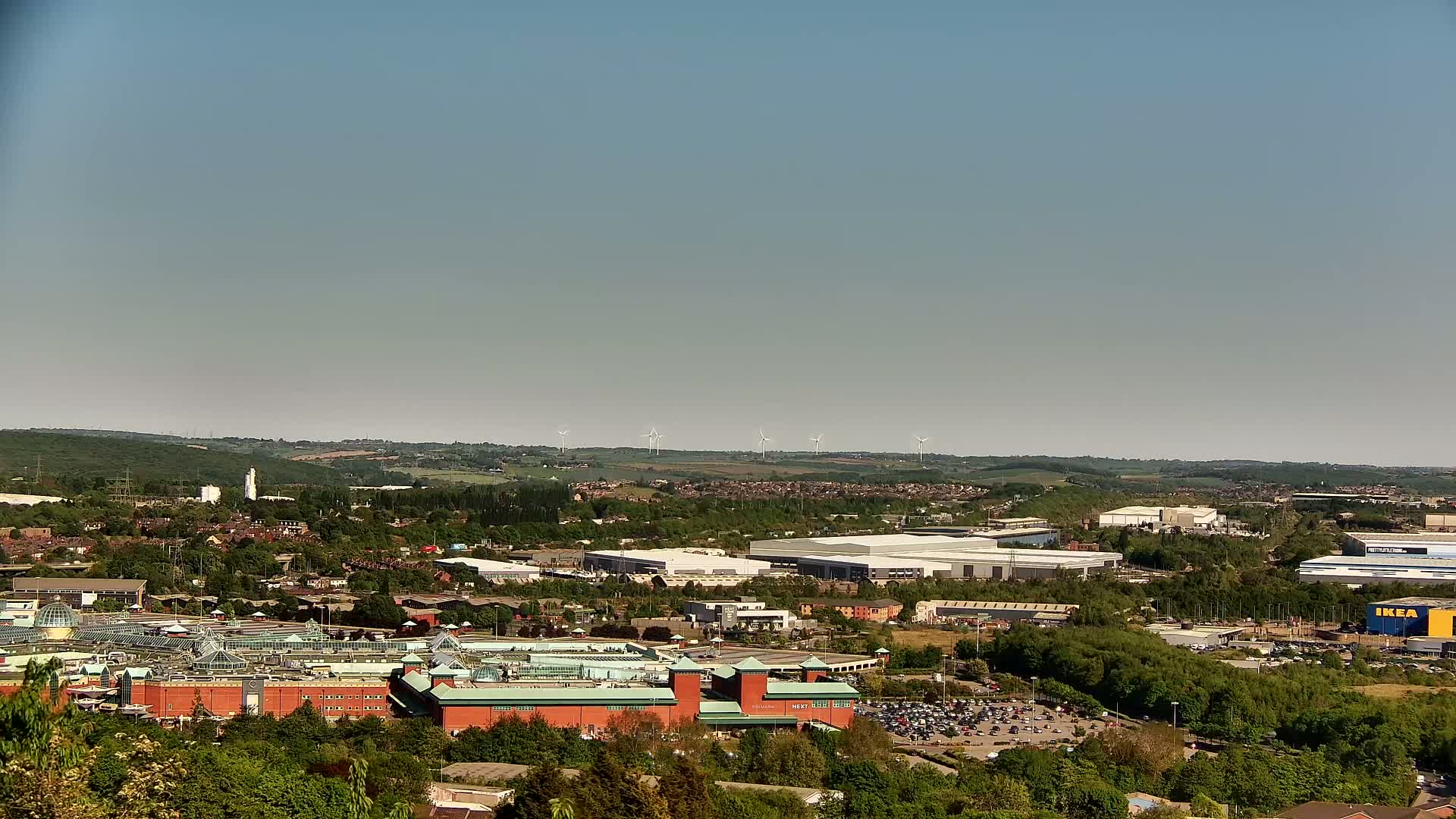 A sprawling suburban landscape with a large shopping mall and several wind turbines visible in the distance under a clear, sunny sky.