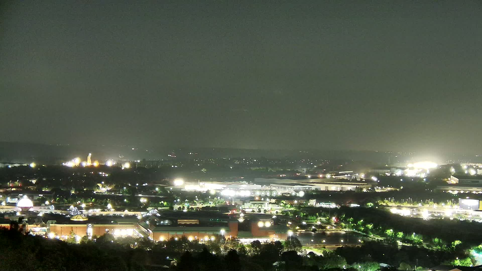 A hazy, night-time view of a city illuminated by numerous lights, featuring a large shopping center and industrial areas nestled amongst trees.