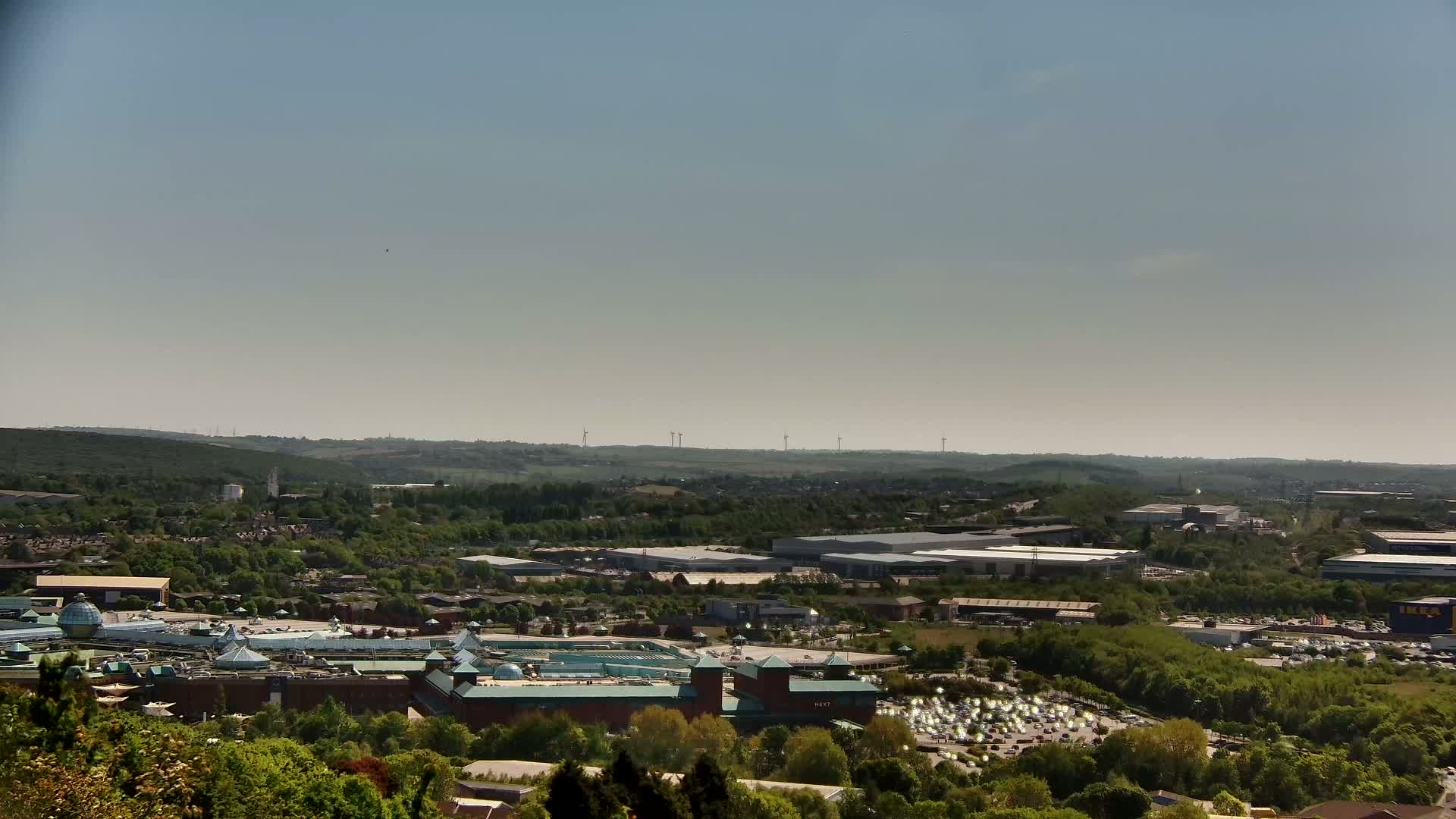 A large shopping mall with a sprawling parking lot is nestled within a green, hilly landscape under a clear, bright sky.