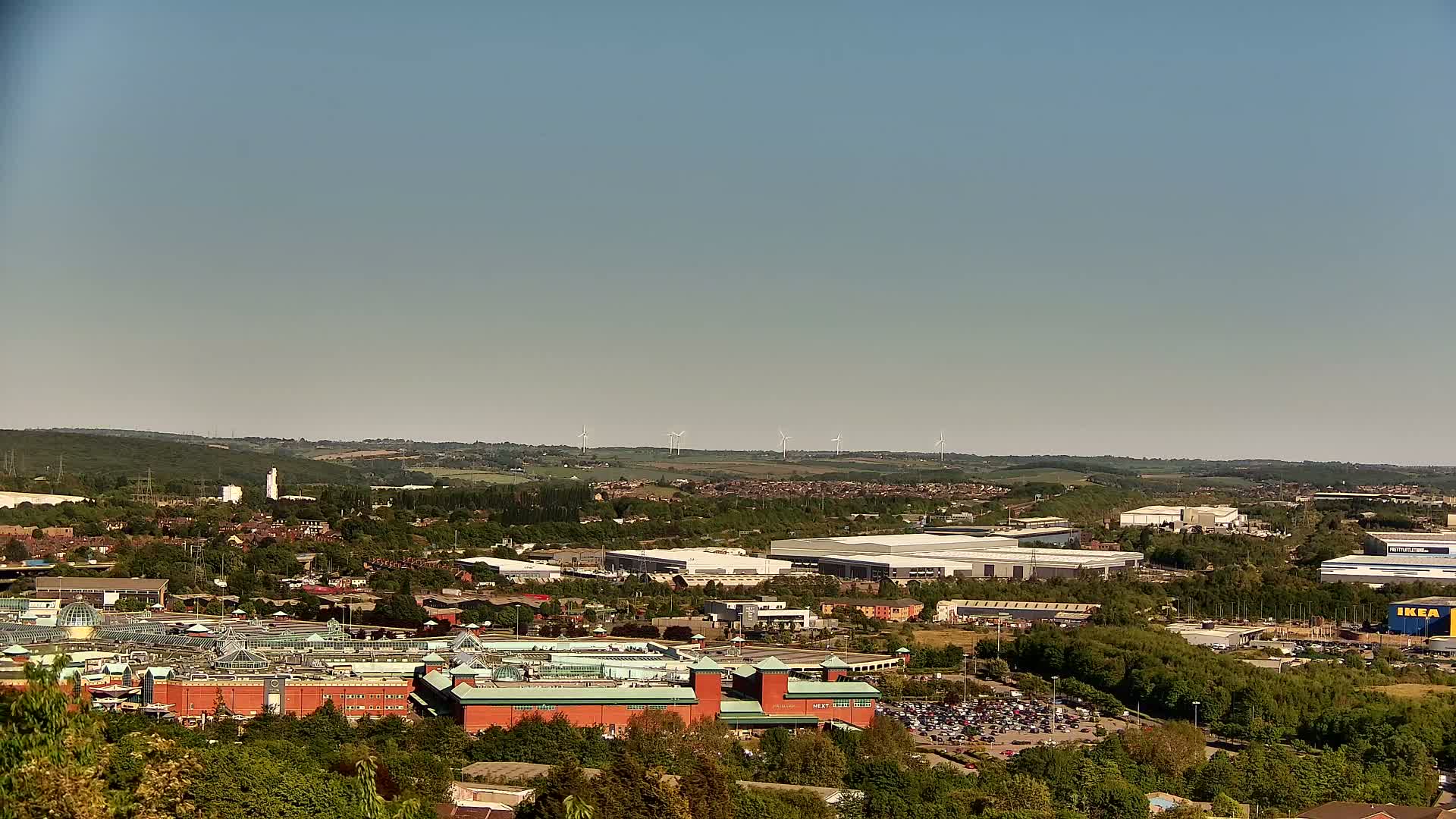 A sprawling suburban landscape under a clear sky features a large shopping mall, industrial buildings, and a line of distant wind turbines.