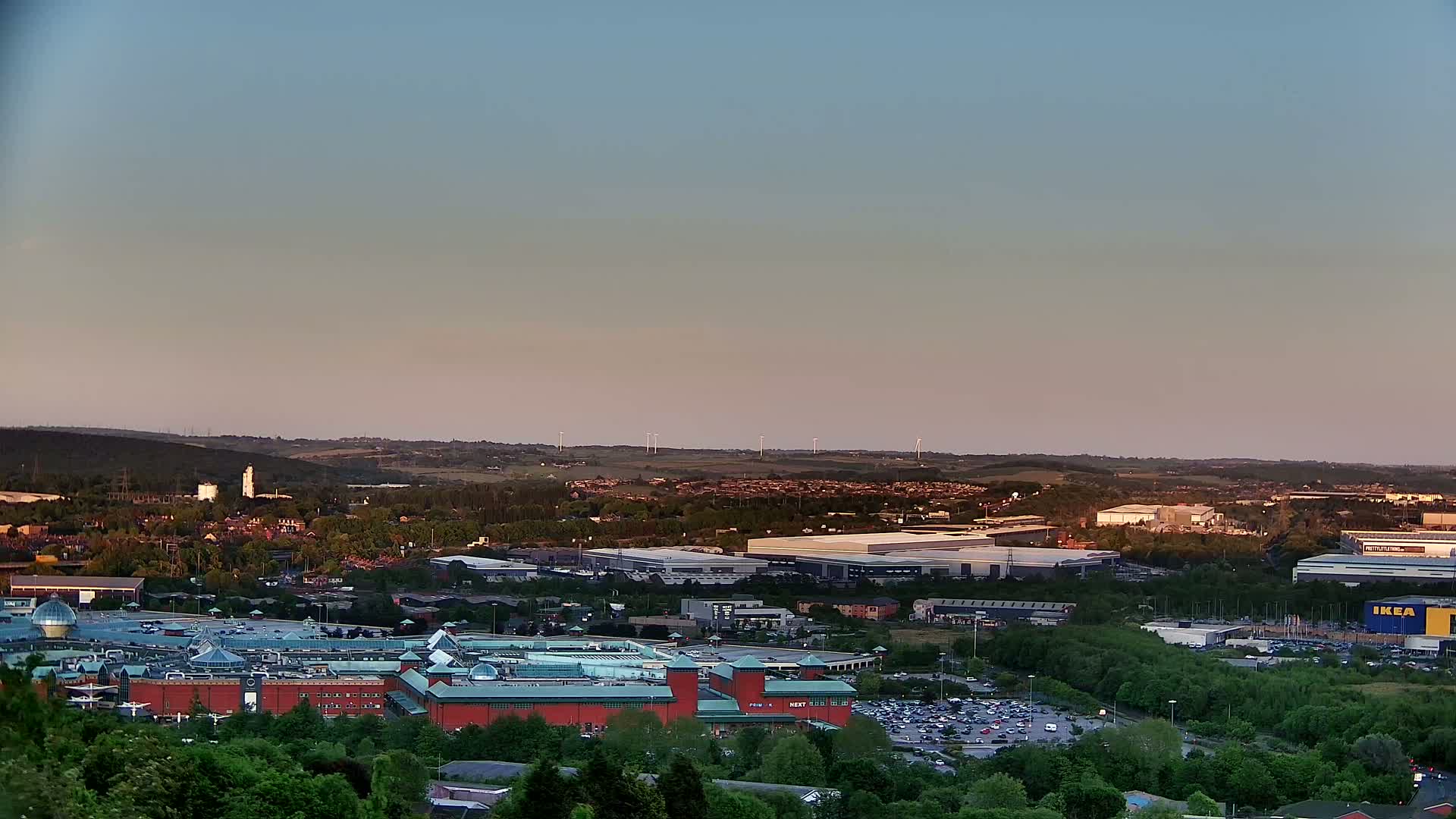 A sprawling suburban landscape at sunset, featuring a large shopping mall and various commercial buildings amidst residential areas and green spaces, with a line of wind turbines visible in the distance.
