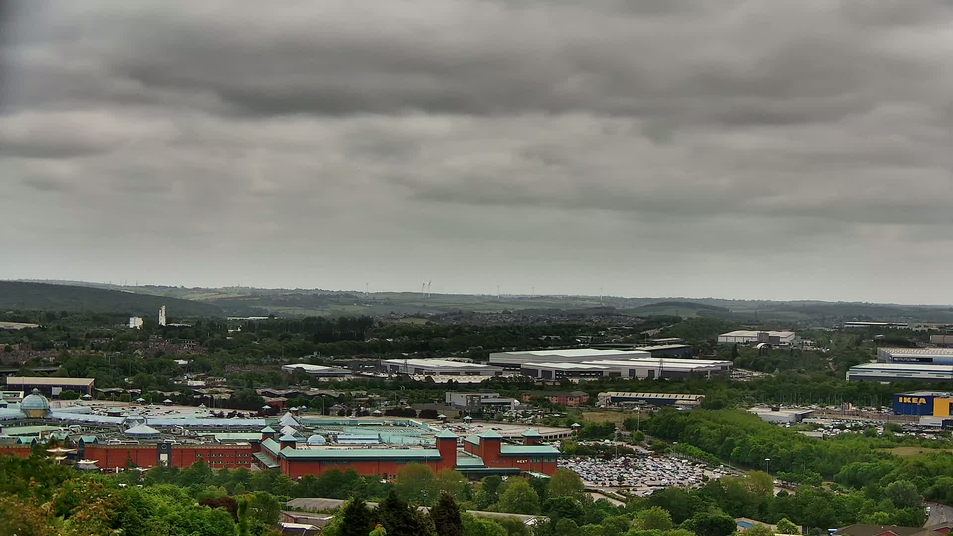 A large shopping mall and surrounding industrial buildings are visible from an elevated viewpoint under a cloudy sky.