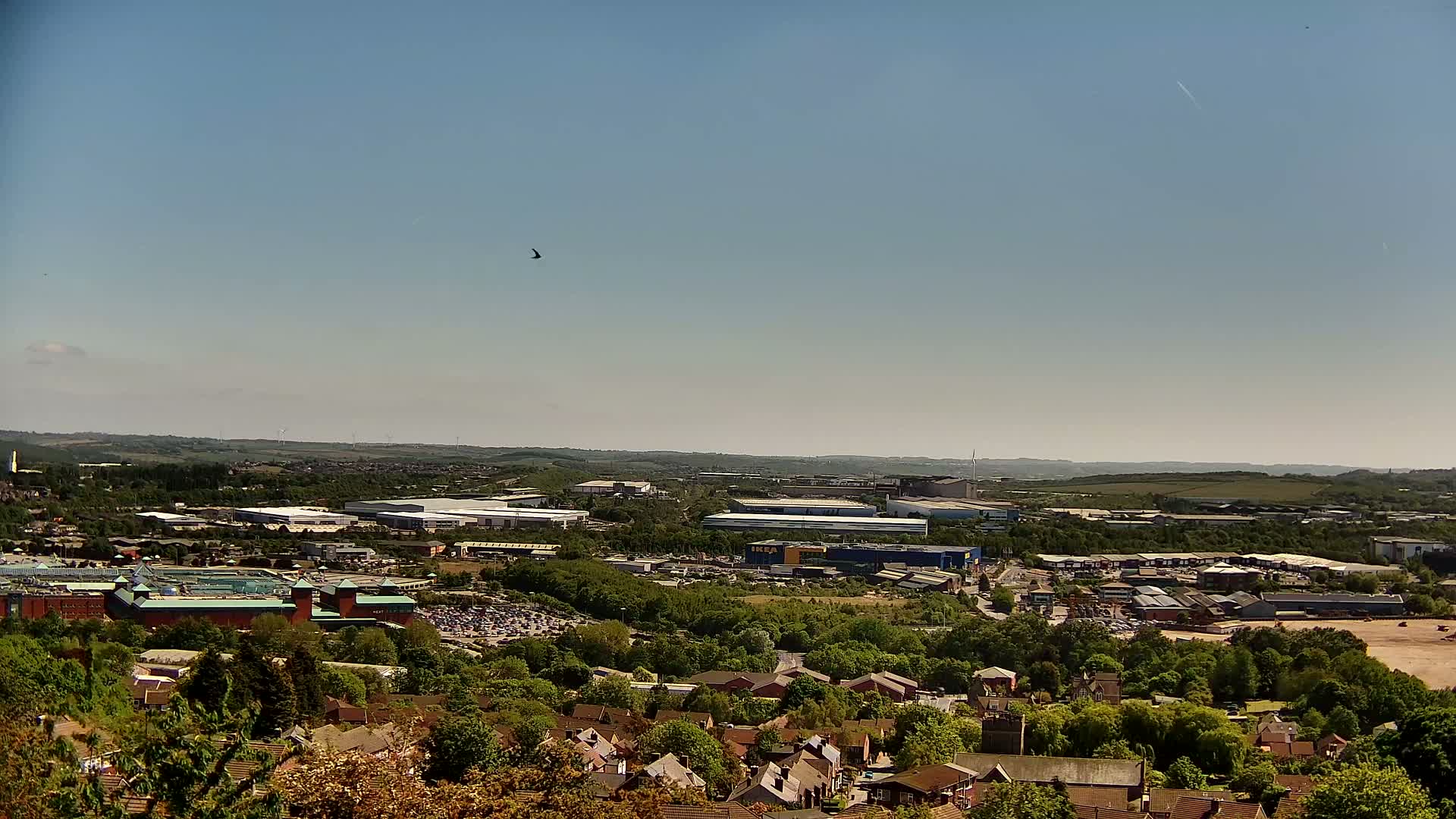 A high-angle, long shot on a clear, sunny day shows a sprawling suburban area with industrial buildings and a large retail complex nestled amongst green trees and houses.