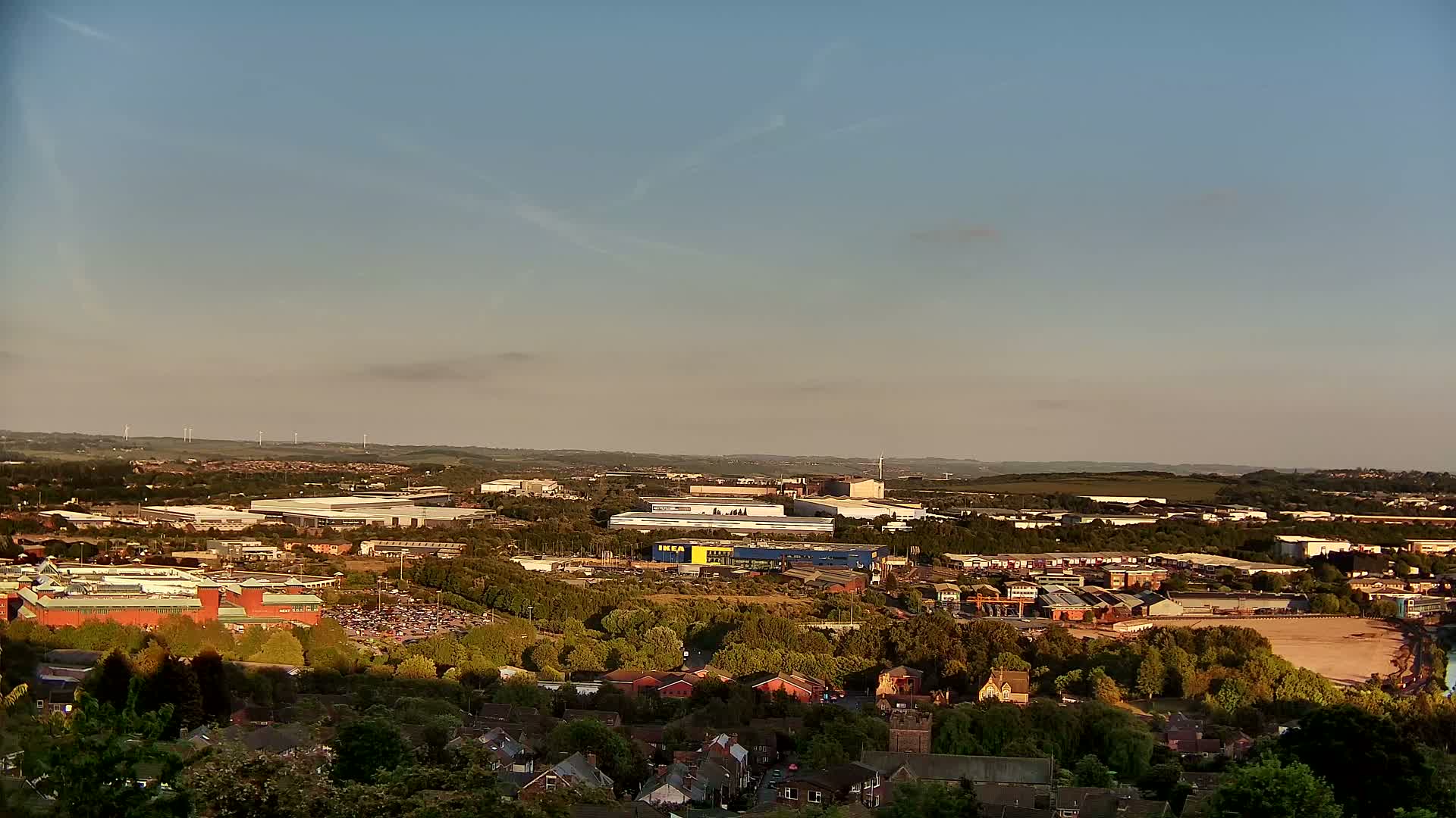 An elevated view of a suburban landscape under a clear, light blue sky shows a large retail park and industrial buildings nestled amongst green trees and houses.