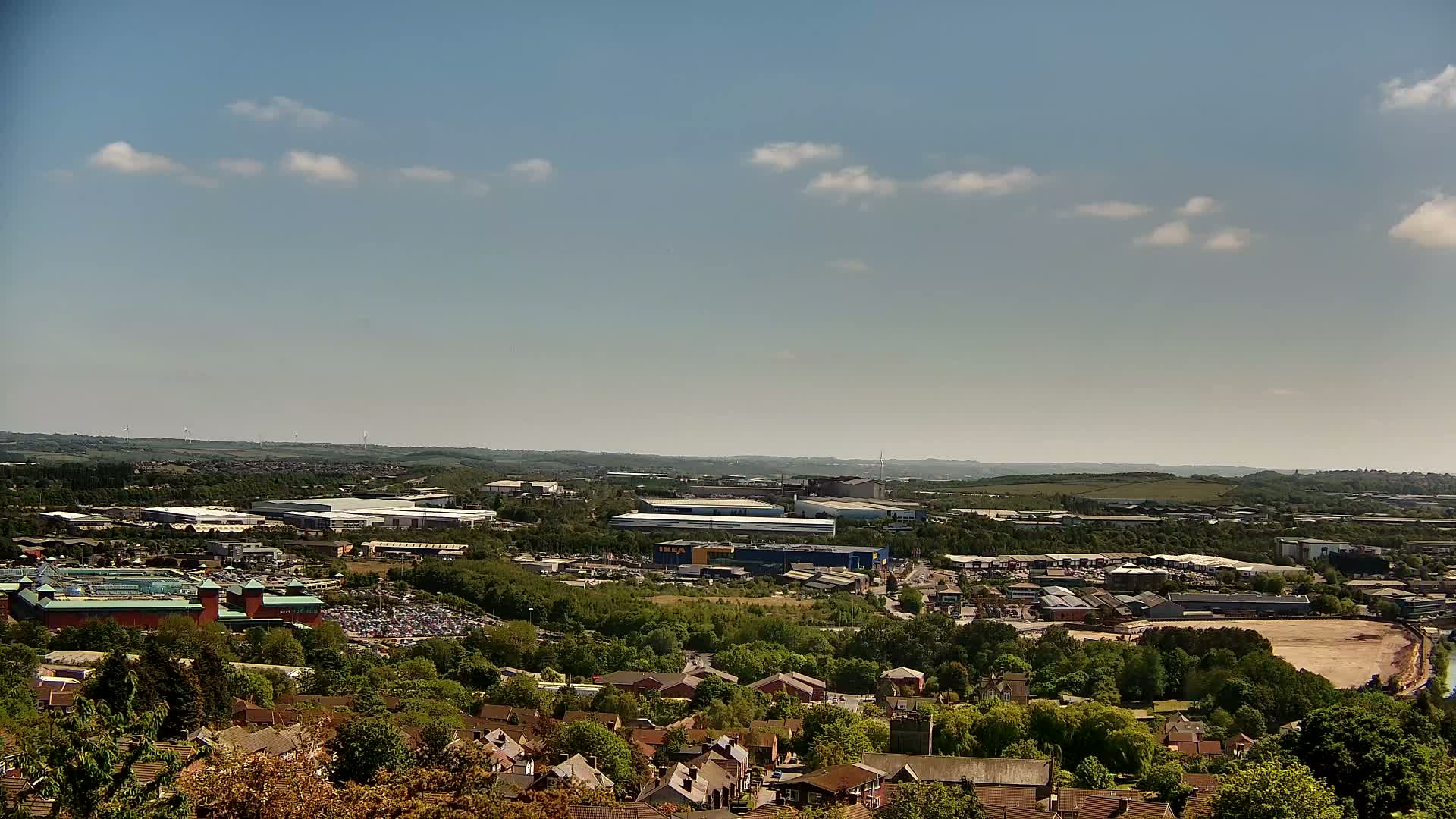 A sprawling suburban landscape under a mostly clear, sunny sky shows a mix of residential homes, industrial buildings, and a large retail complex.