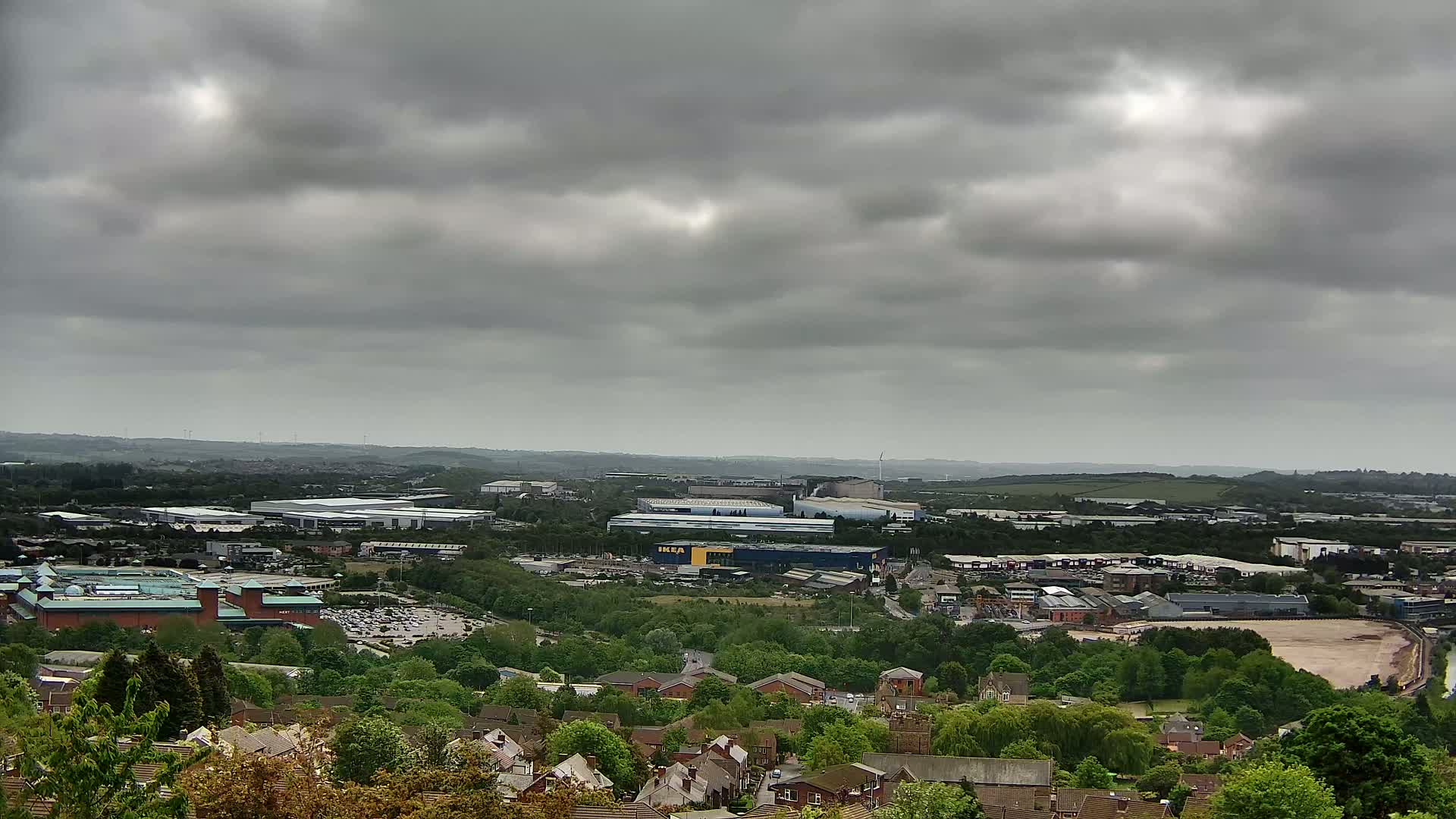 A sprawling suburban landscape under a heavily overcast sky features a large shopping center and numerous buildings, interspersed with green spaces and residential areas.