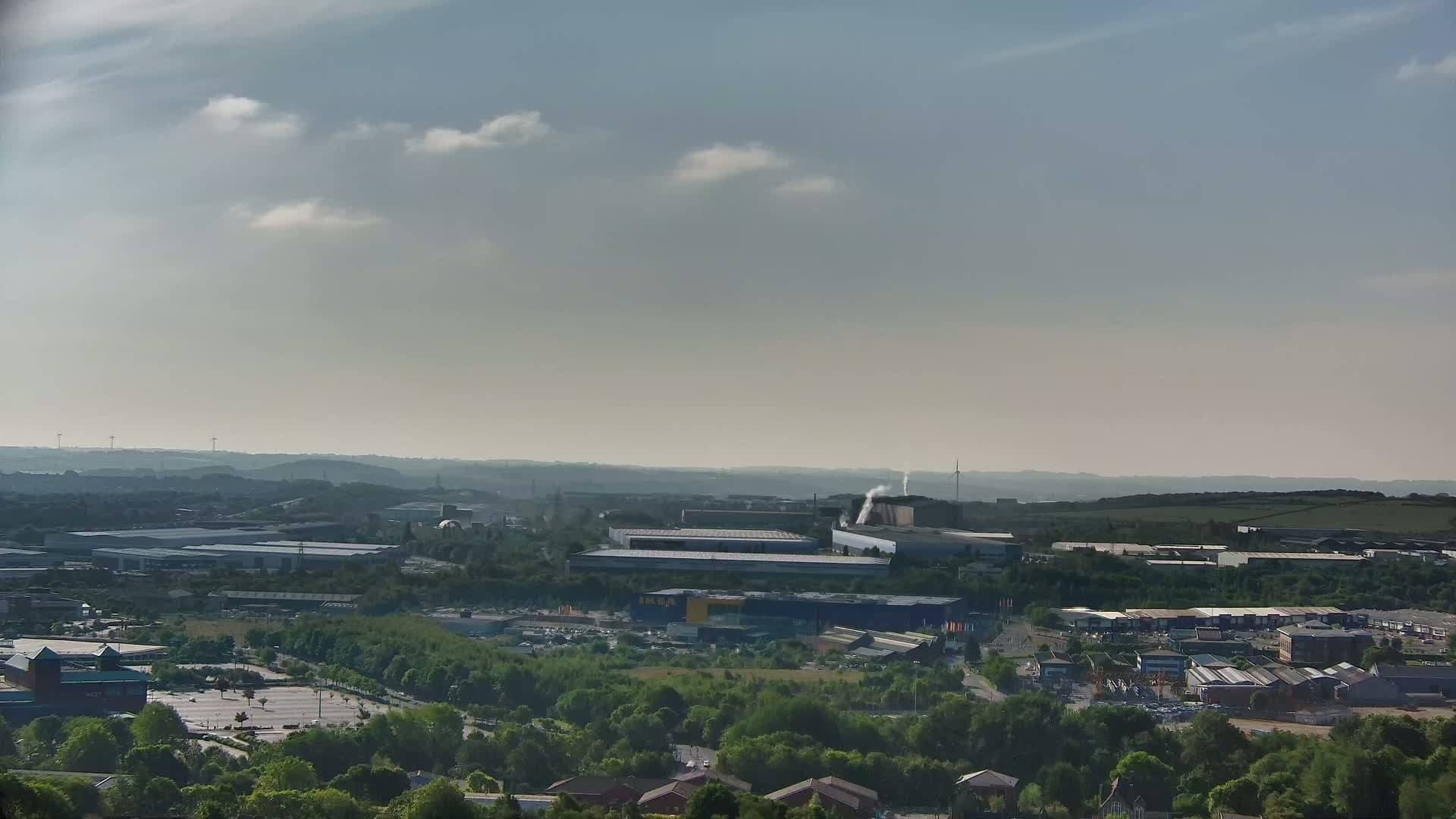 A hazy, sunny day reveals a sprawling industrial area dotted with warehouses and buildings nestled among green trees and a parking lot, viewed from an elevated vantage point.