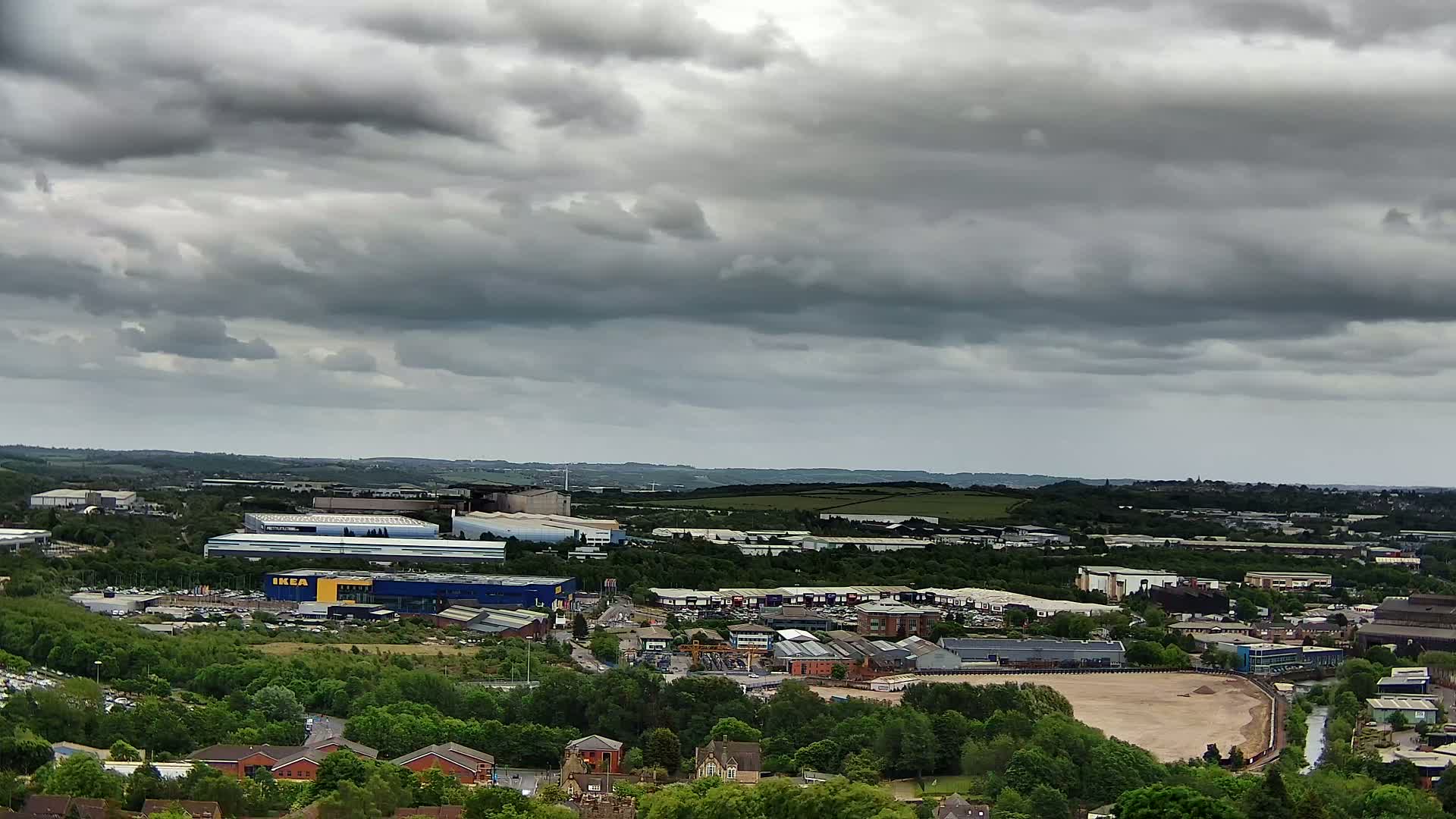 An expansive view of an industrial area, dotted with buildings and surrounded by lush green trees, under a heavily clouded sky.