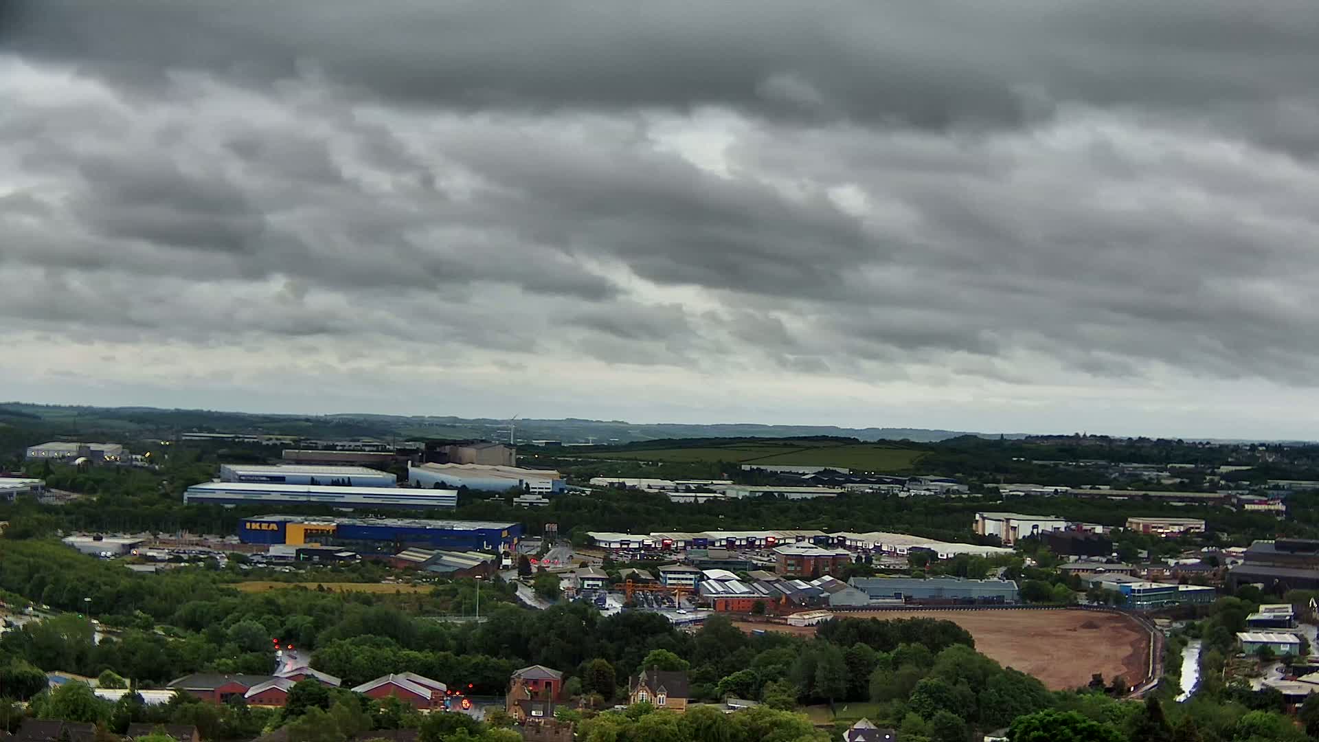 A high-angle view of an industrial area with overcast skies shows a large complex of buildings and warehouses, surrounded by green trees and a cleared, reddish-brown area of land.