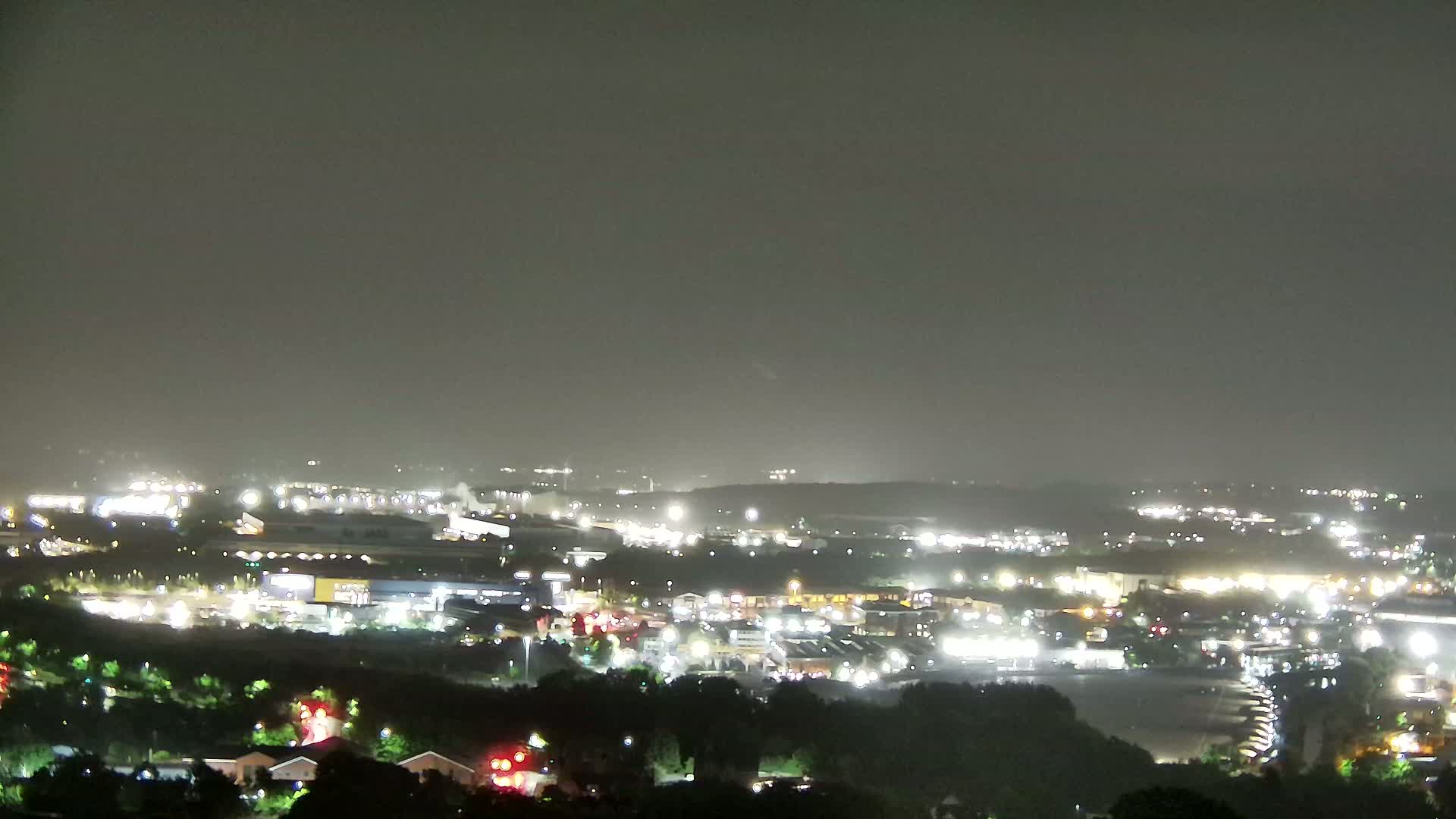 A nighttime view of a city landscape under a hazy sky, showing numerous illuminated buildings and structures spread across a valley, surrounded by dark vegetation.