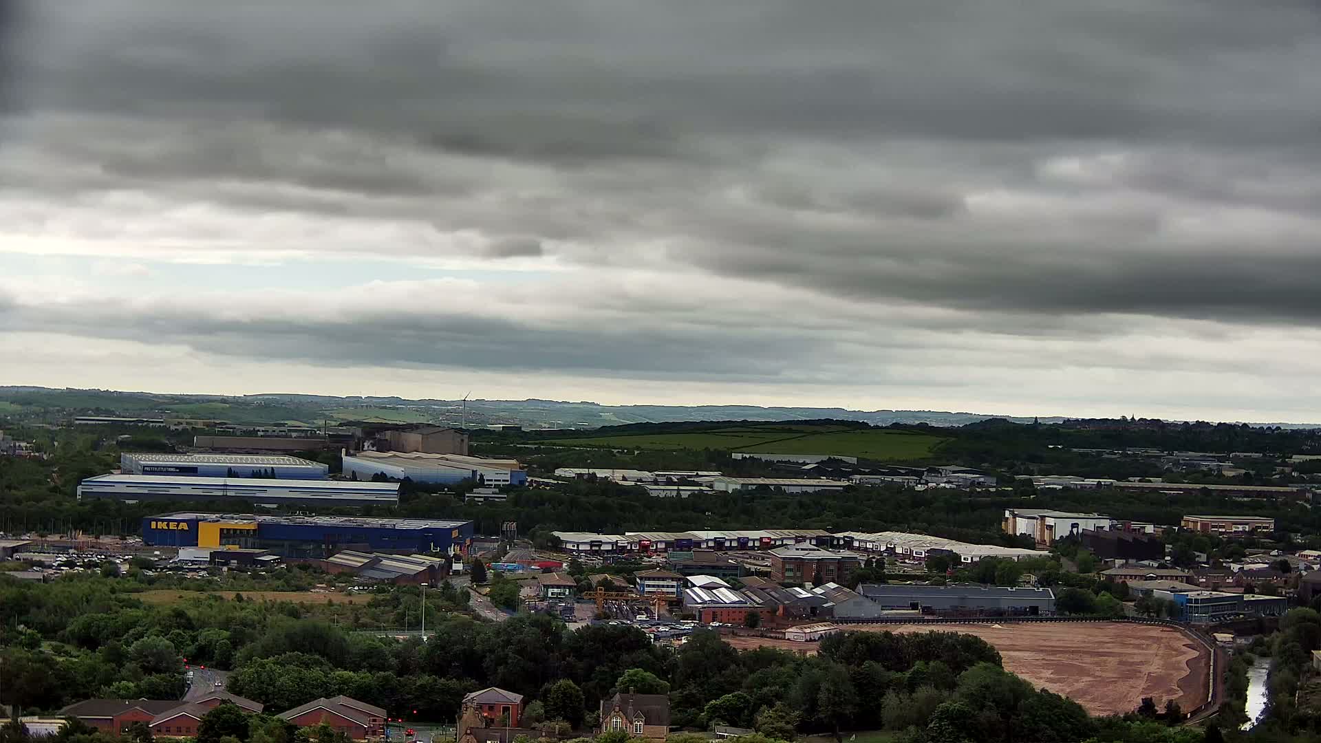 An expansive view of an industrial area and surrounding landscape under a heavily overcast sky shows a large, flat expanse of exposed earth, surrounded by green trees and buildings, with an IKEA store prominently featured among numerous other structures.