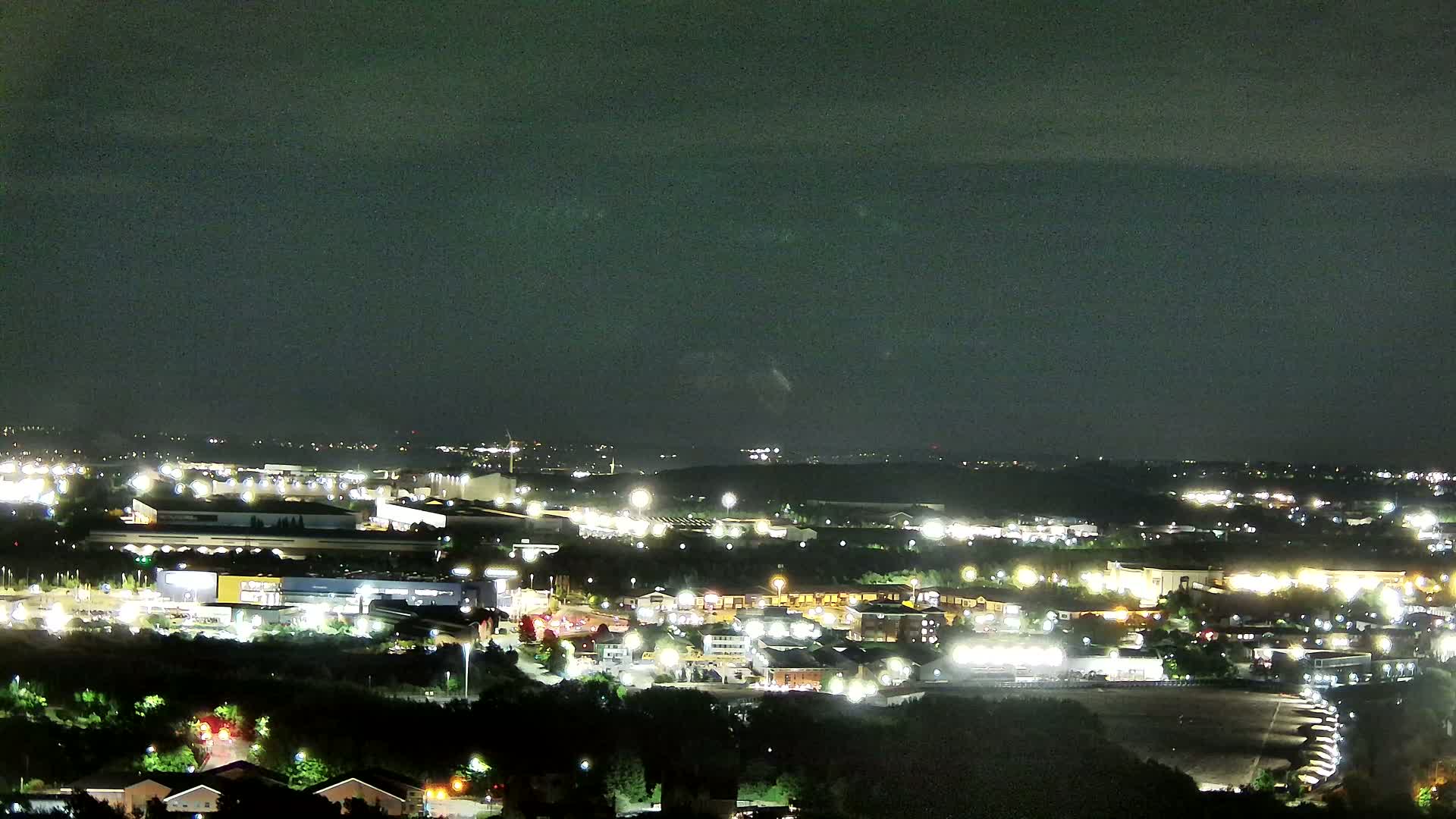 A nighttime, high-angle view shows a sprawling suburban area with numerous lit buildings and roadways under a dark, cloudy sky.