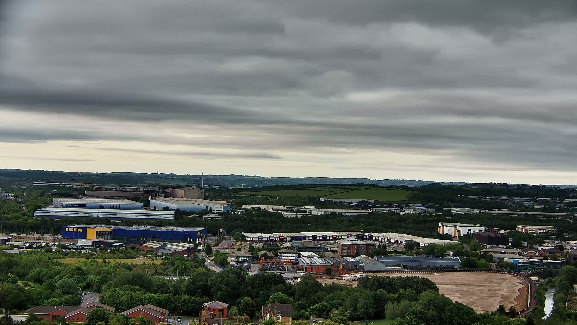 An aerial view of an industrial area with a large blue and yellow building, surrounded by greenery and under a cloudy sky.