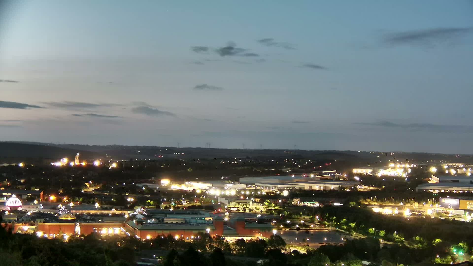 A nighttime view of a sprawling illuminated city, featuring a large shopping mall and surrounding buildings under a twilight sky with a few clouds.