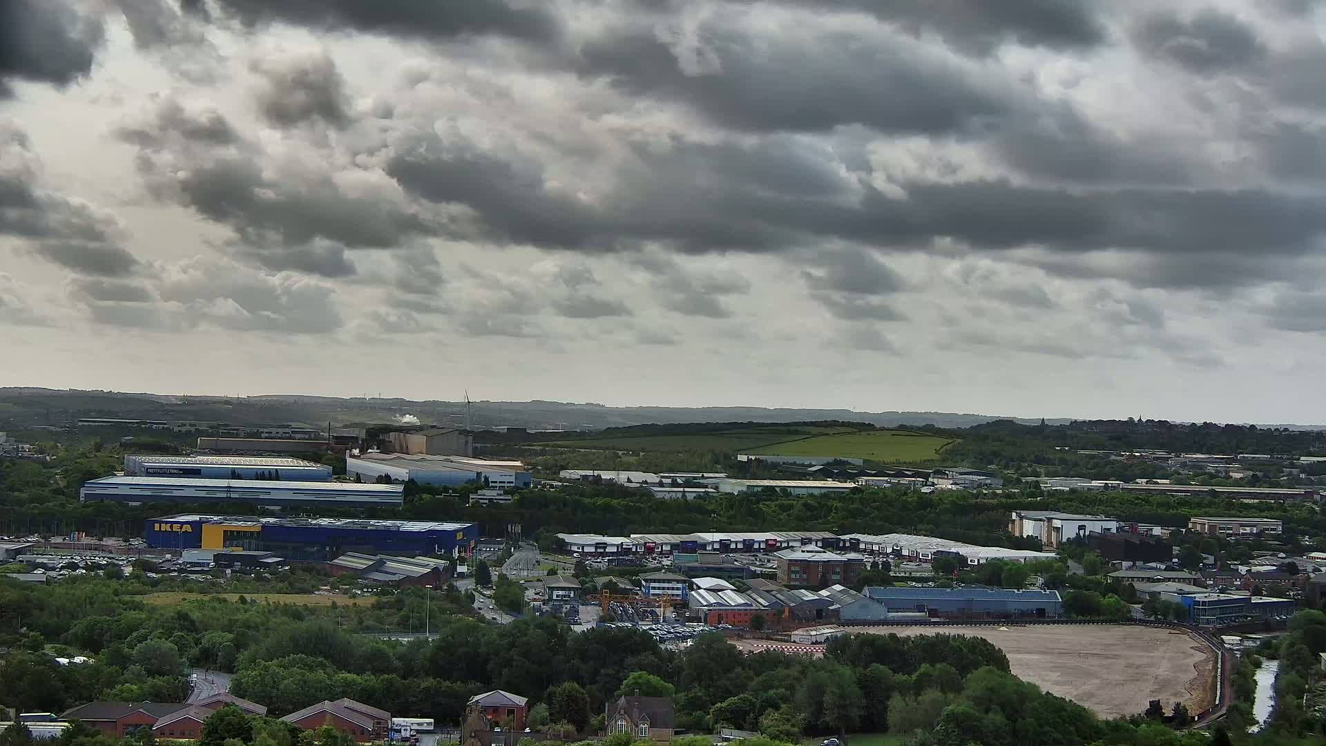 An elevated view of an industrial and commercial area under a cloudy sky shows a mix of large warehouses and smaller buildings, surrounded by green trees and a large empty field.