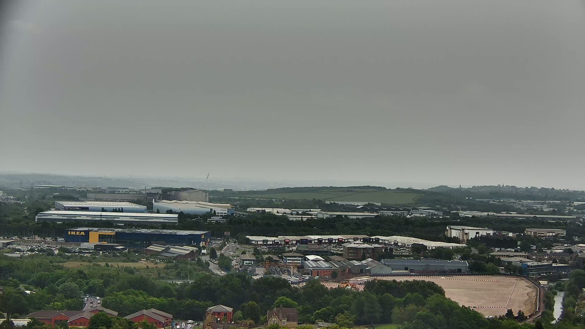 An aerial view of an industrial area on an overcast day, showing various buildings including a large blue-and-yellow structure, surrounded by greenery and a cleared, reddish-brown field.