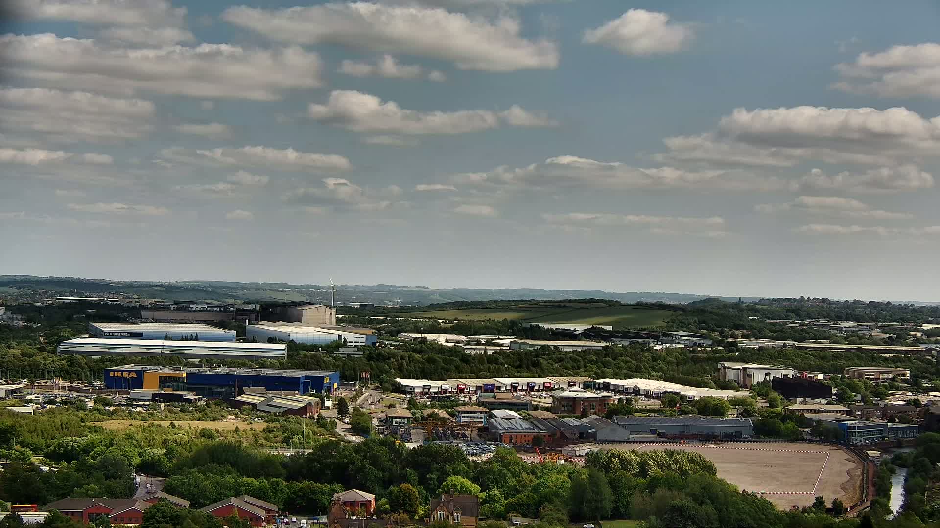 A high-angle view of an industrial area with a large blue building, other commercial structures, and surrounding green spaces under a partly cloudy sky.
