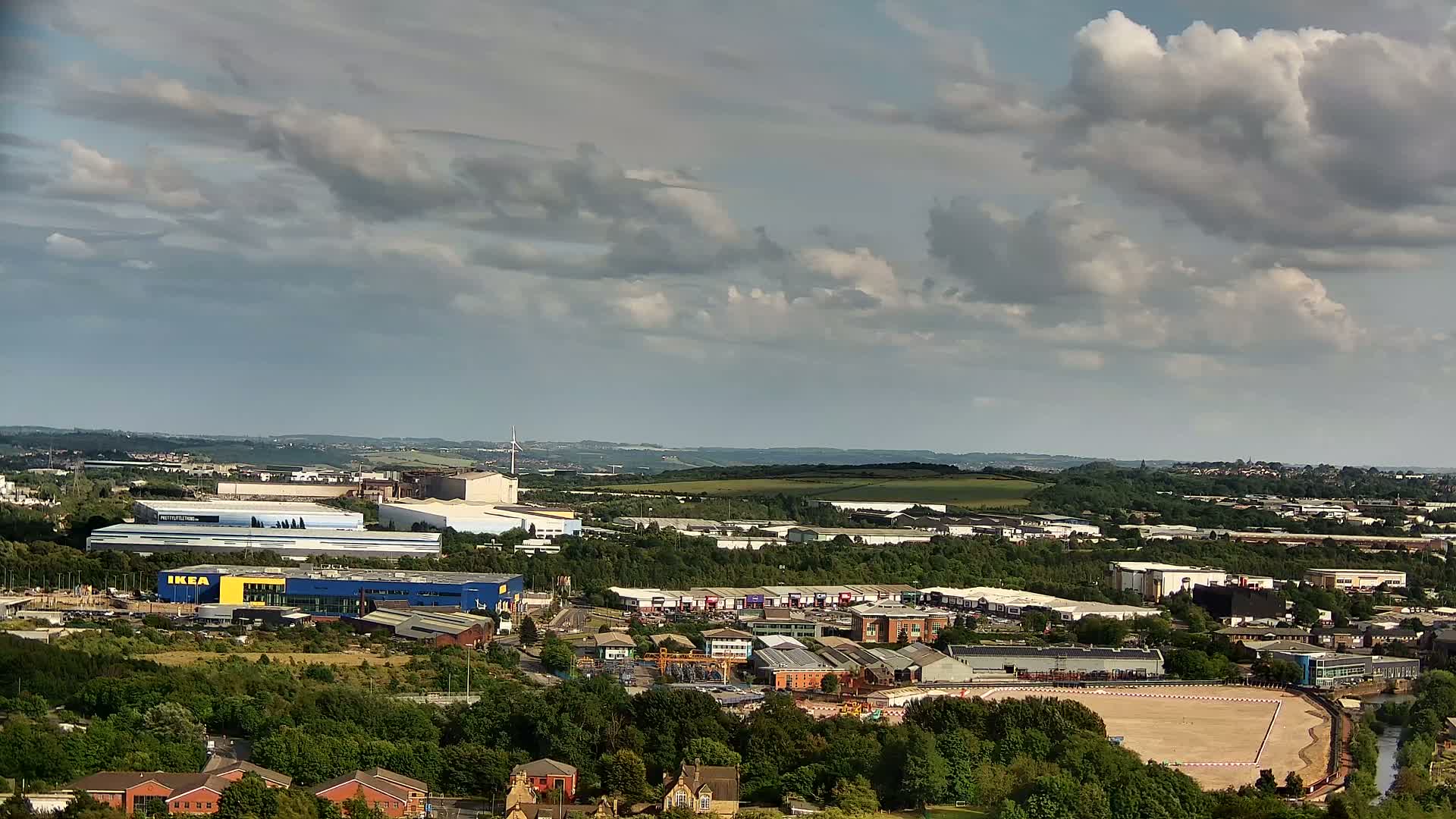 A sprawling industrial and residential area under a partly cloudy sky, featuring a large blue-and-yellow building among numerous other structures and a cleared, sandy area.