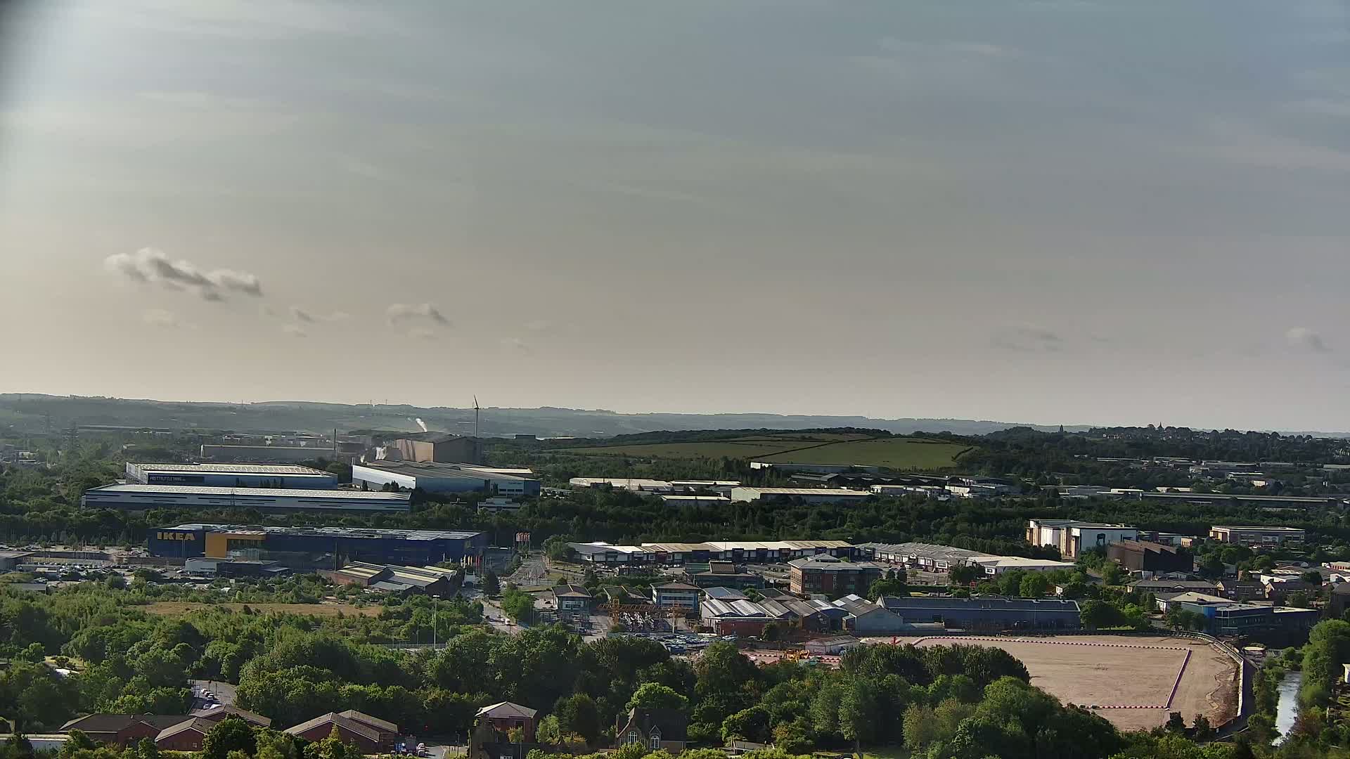 An expansive view of an industrial and commercial area, under a partly cloudy sky, shows a mix of buildings, green spaces, and a large empty lot.