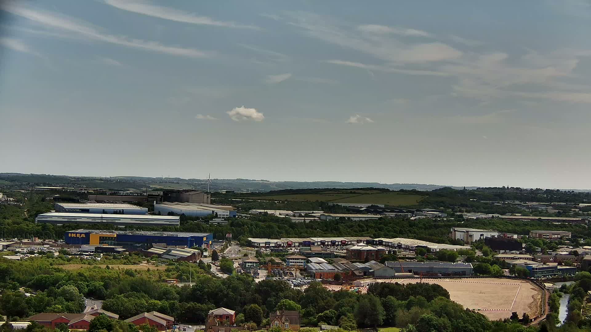 A high-angle, long shot on a partly sunny day shows an industrial area with an IKEA store, surrounded by greenery and other buildings, set against a backdrop of rolling hills.