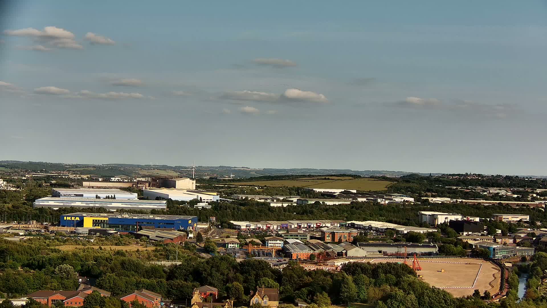 An aerial view shows a sprawling industrial area with an IKEA store, surrounded by green trees and fields under a partly cloudy sky.