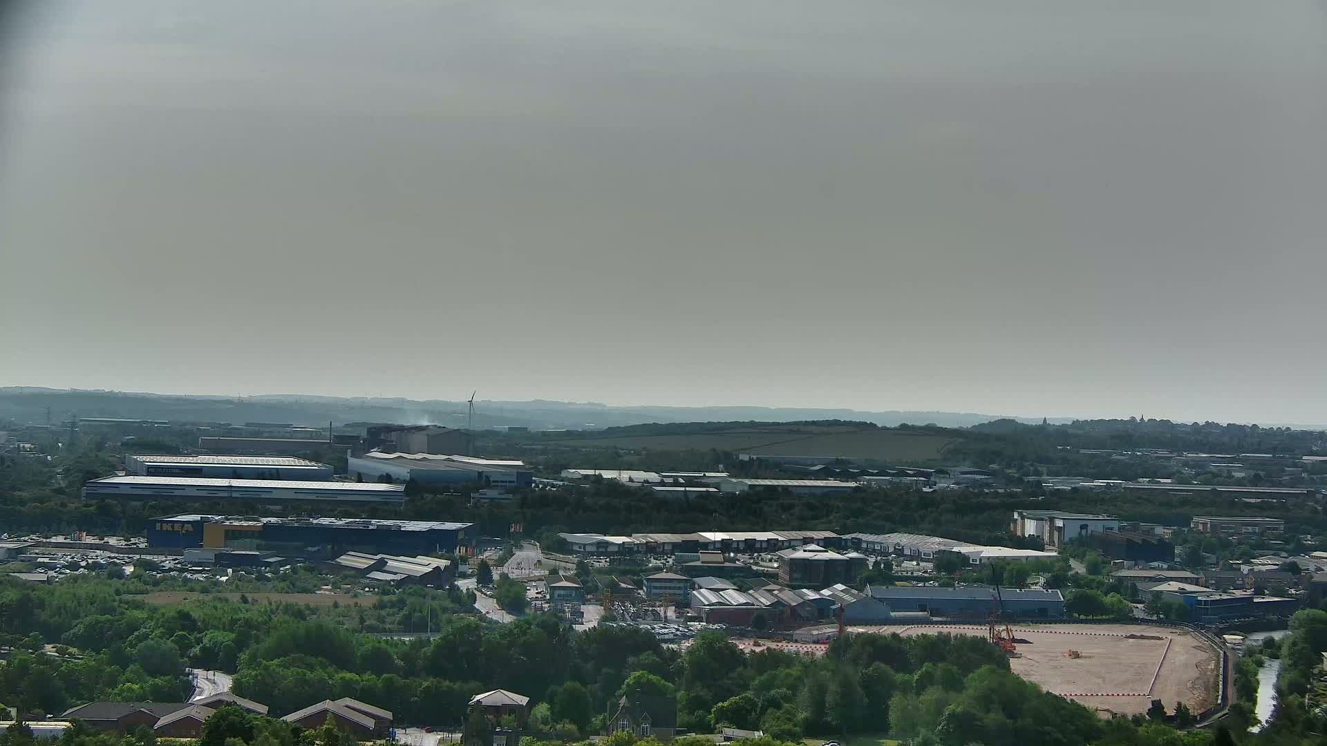 An aerial view shows a hazy, sunny day over a sprawling industrial park and surrounding green landscape, with a large cleared area of land visible in the foreground.