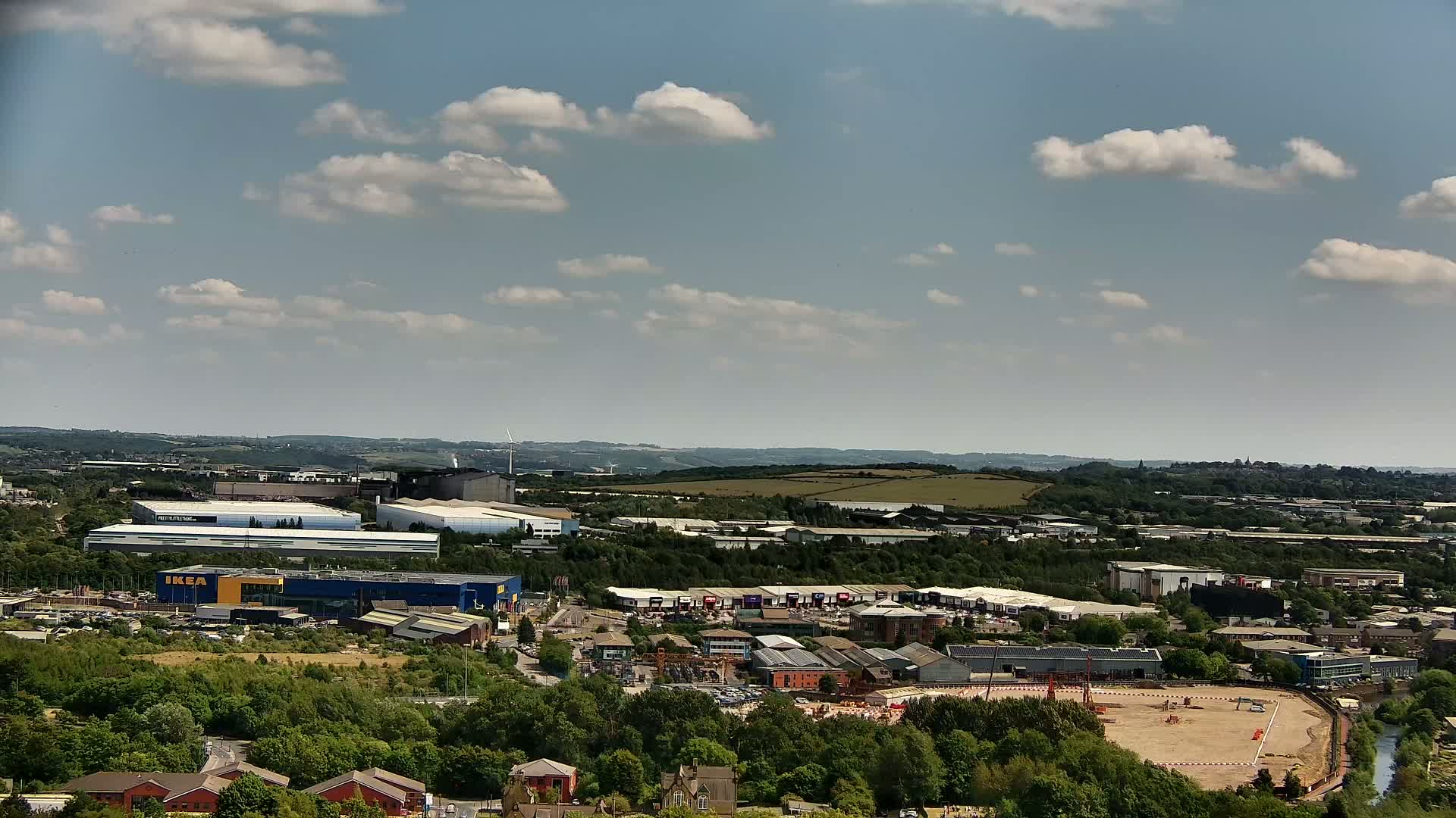 An expansive view of an industrial area with a large blue building, a cleared construction site, and a surrounding landscape under a partly cloudy sky.