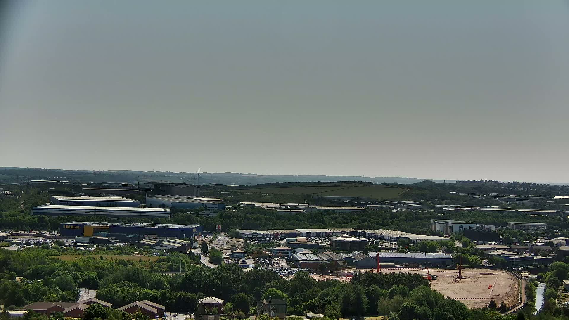 An aerial view shows a large industrial and retail park on a sunny day, with green hills and trees in the background.