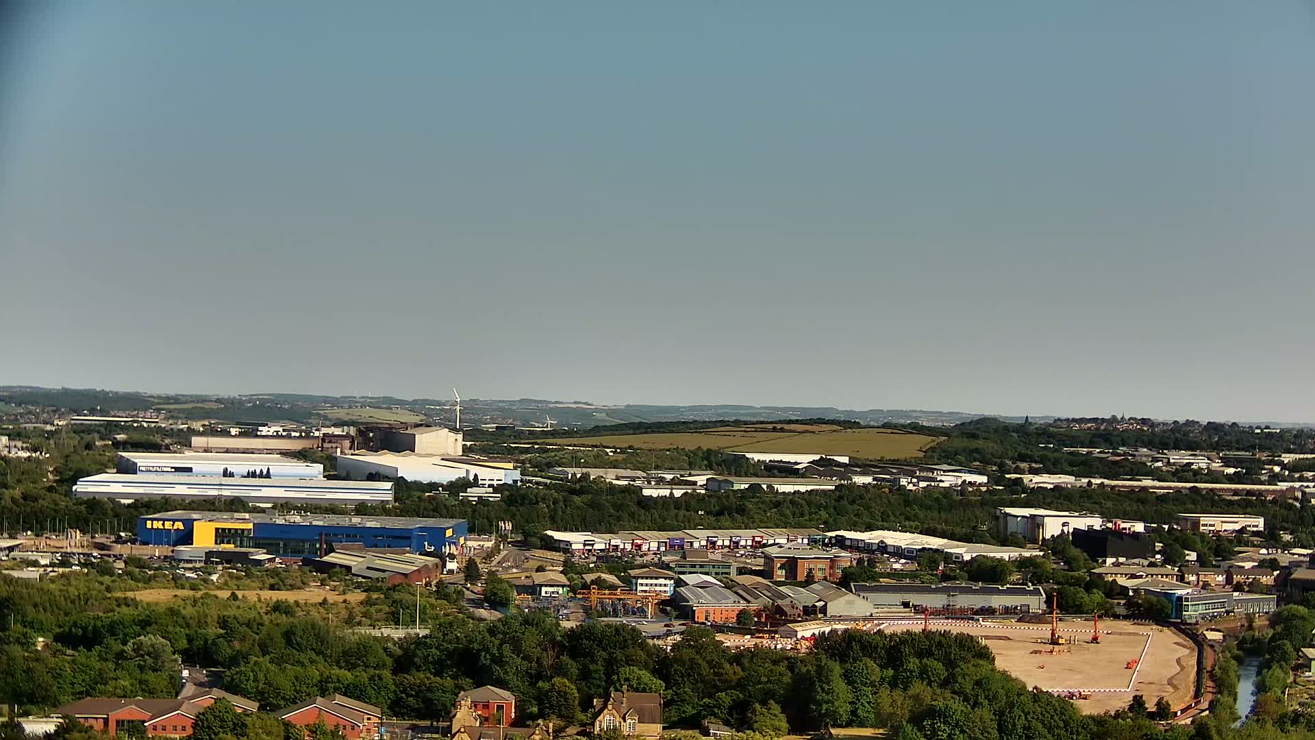 An aerial view shows a large industrial area with various buildings, including a prominent blue and yellow building, under a clear, sunny sky.