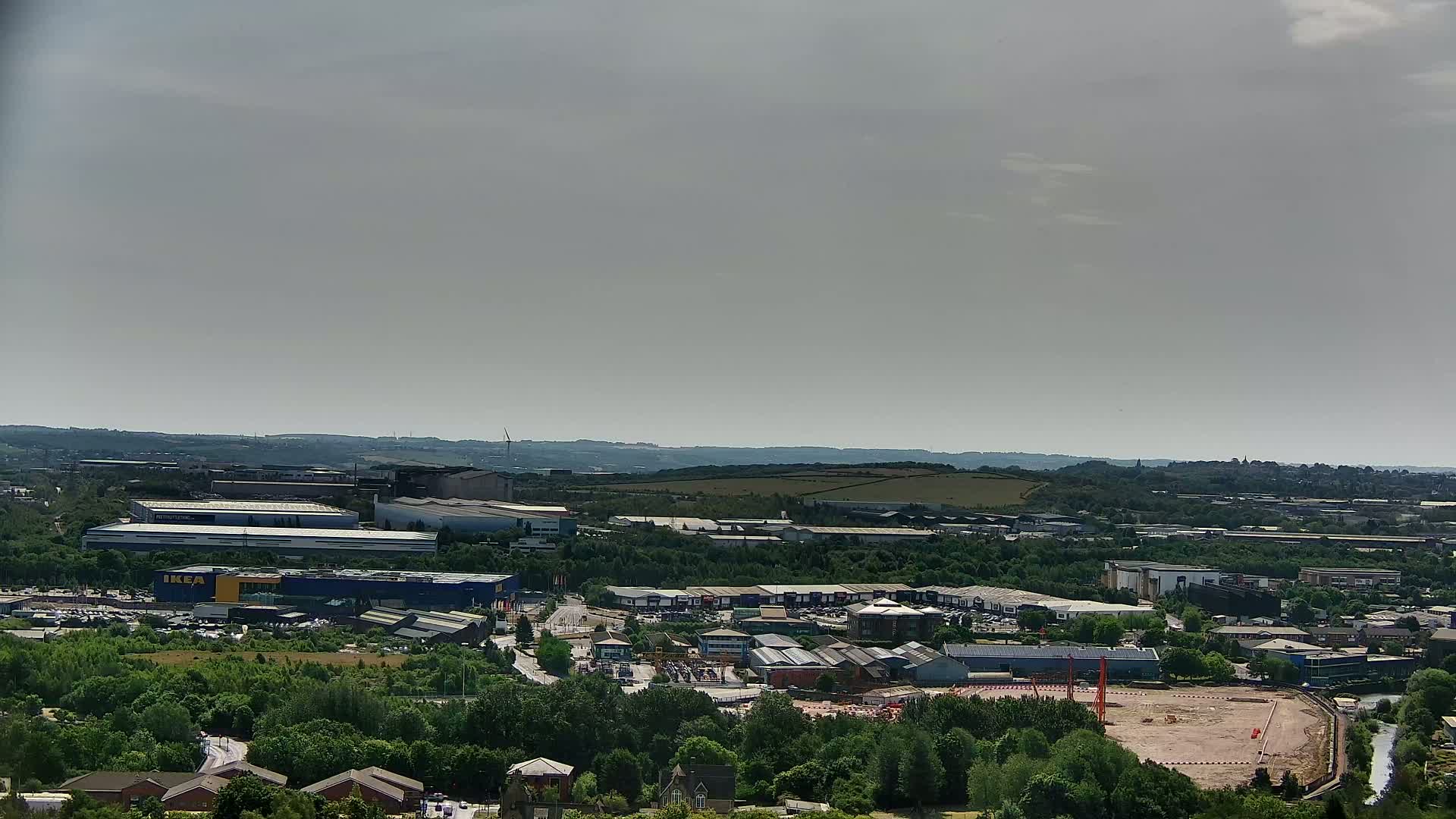 An aerial view shows a sprawling industrial park and retail area nestled among green trees and hills under a hazy sky.