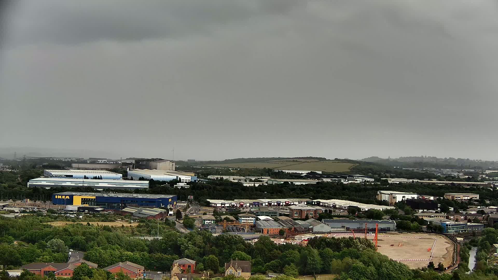 An overcast day reveals an aerial view of an industrial area with various buildings, including a large blue structure, surrounded by greenery and a cleared construction site.