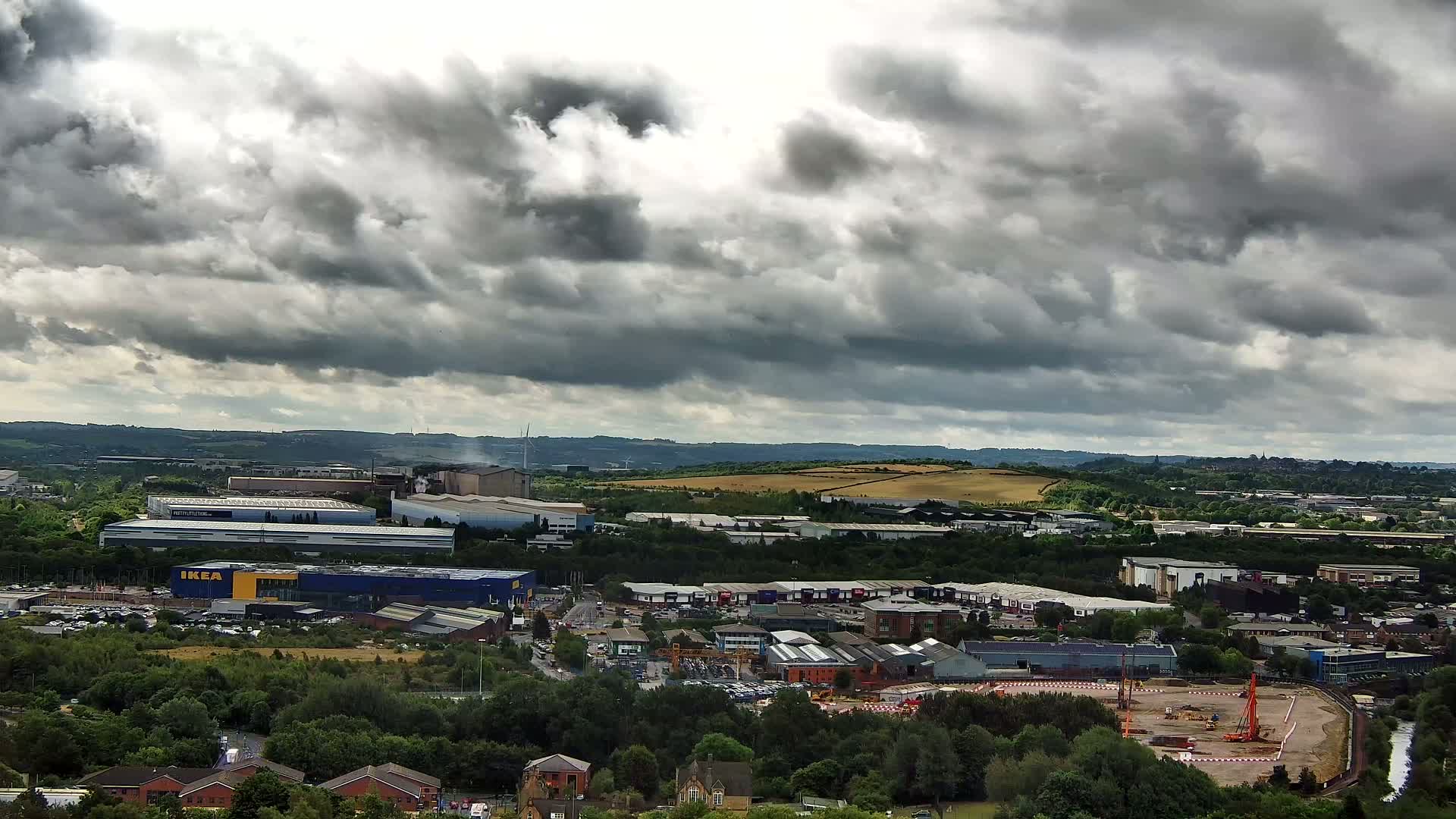 An expansive view of an industrial area with a large blue building, surrounded by greenery and under a cloudy sky.