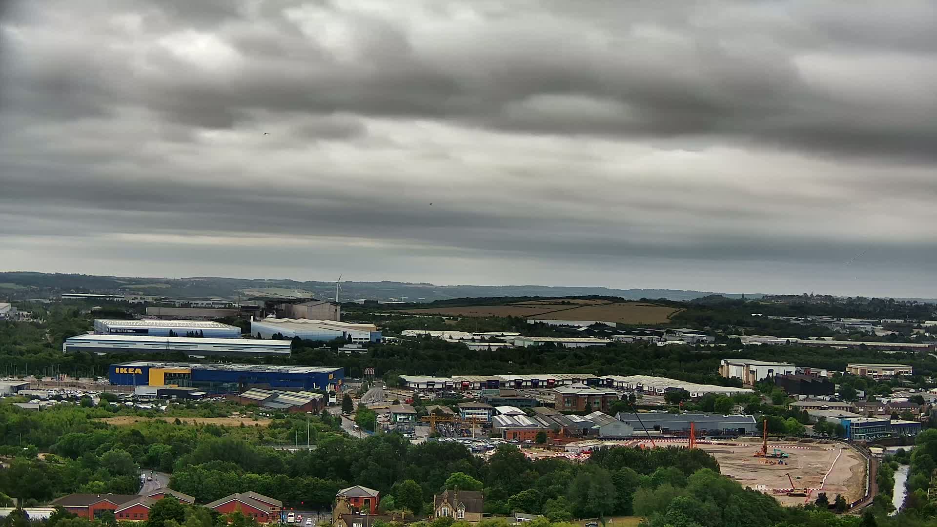 An expansive view of an industrial area and retail park under a cloudy sky, with a large construction site visible in the foreground.