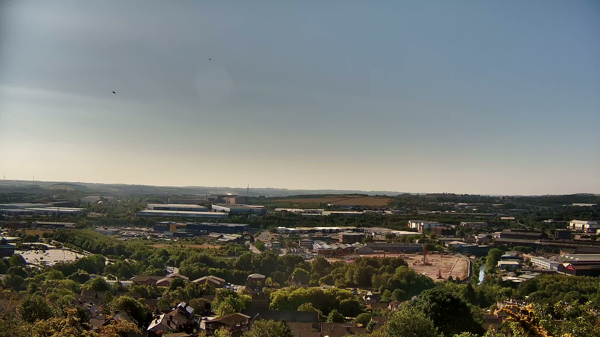 A high-angle, long shot on a clear, sunny day shows a sprawling industrial area with numerous buildings and warehouses interspersed with green spaces and residential areas.