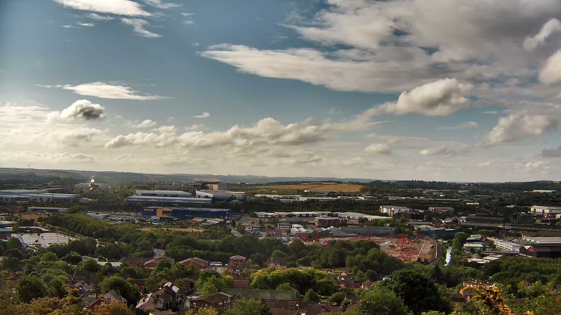 A high-angle view of a partly cloudy day shows an industrial area and residential neighborhood nestled among green trees, with a distant landscape under a partly cloudy sky.