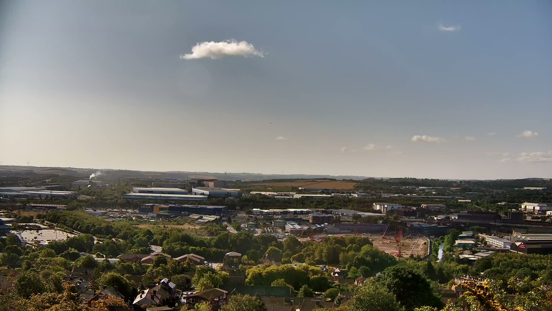 A high-angle view shows a sprawling industrial area and residential neighborhood nestled amongst green trees under a mostly clear sky with a few scattered clouds.