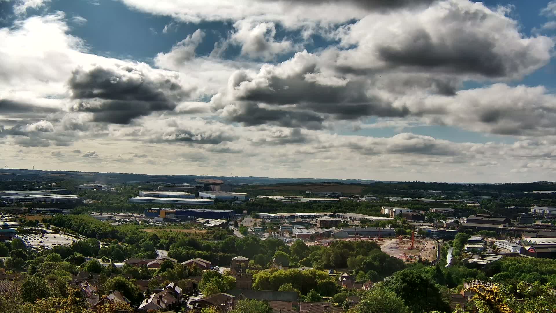 A sprawling landscape under a partly cloudy sky shows a mix of residential homes, industrial buildings, and green spaces.