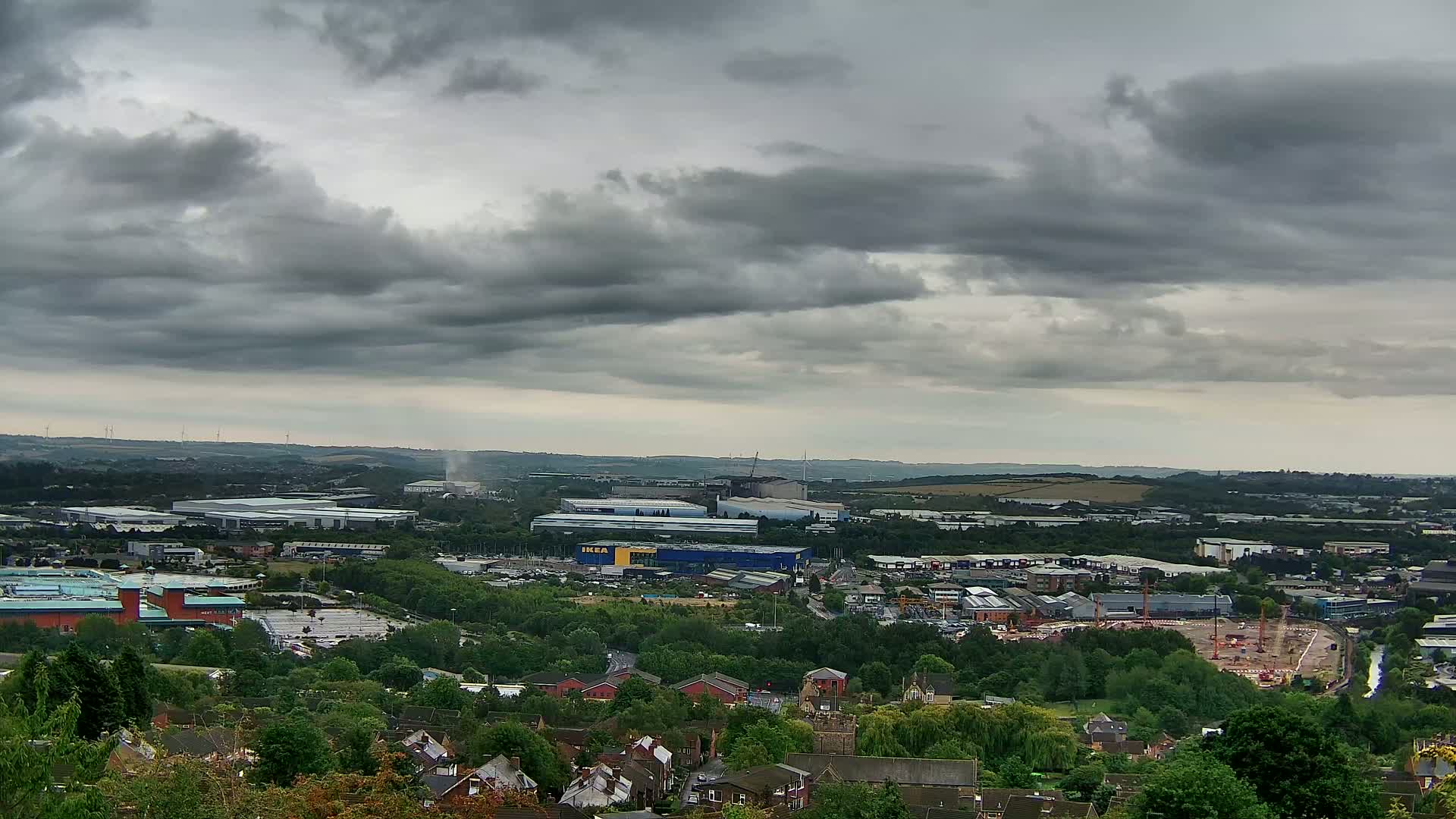 An expansive view of a suburban landscape under a cloudy sky shows industrial buildings, residential areas, and a construction site nestled among green trees.