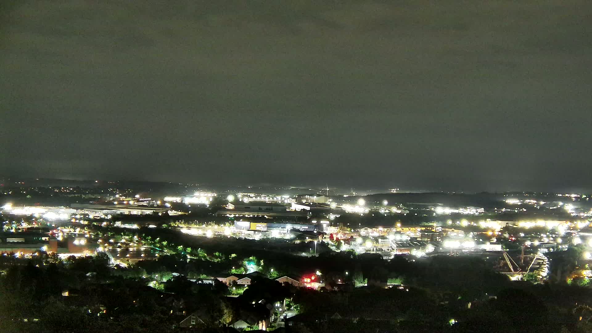 An expansive view of a suburban landscape under a cloudy sky shows industrial buildings, residential areas, and a construction site nestled among green trees.