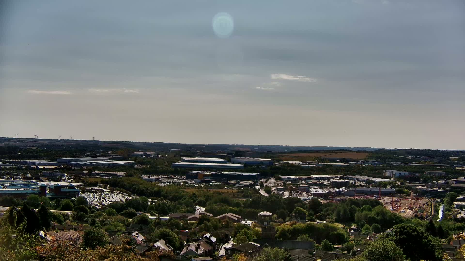 An expansive view of a suburban landscape under a cloudy sky shows industrial buildings, residential areas, and a construction site nestled among green trees.