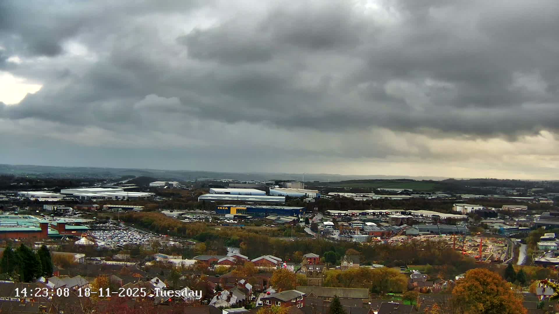 An expansive view of a suburban landscape under a cloudy sky shows industrial buildings, residential areas, and a construction site nestled among green trees.