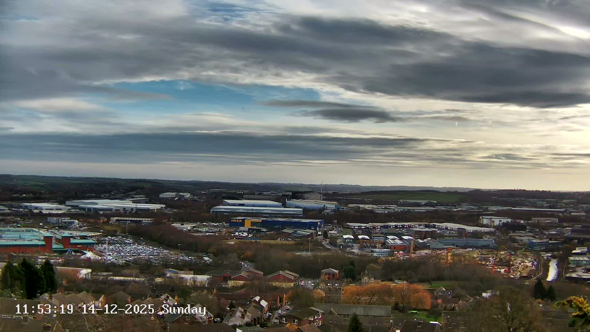 A wide view captures a vast urban and industrial landscape at sunrise, with the sky displaying a mix of blue, grey, and golden-orange clouds, as factories emit steam into the clear morning air.