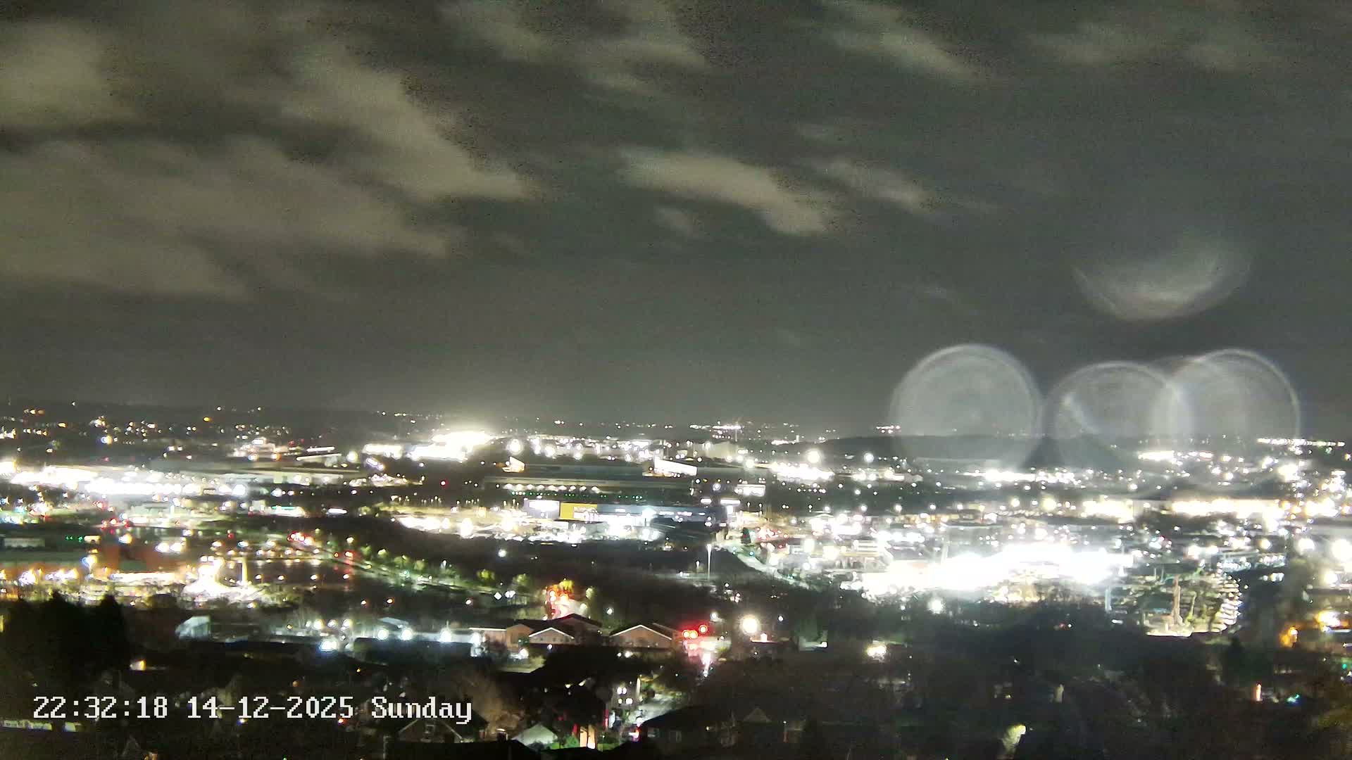 A wide view captures a vast urban and industrial landscape at sunrise, with the sky displaying a mix of blue, grey, and golden-orange clouds, as factories emit steam into the clear morning air.
