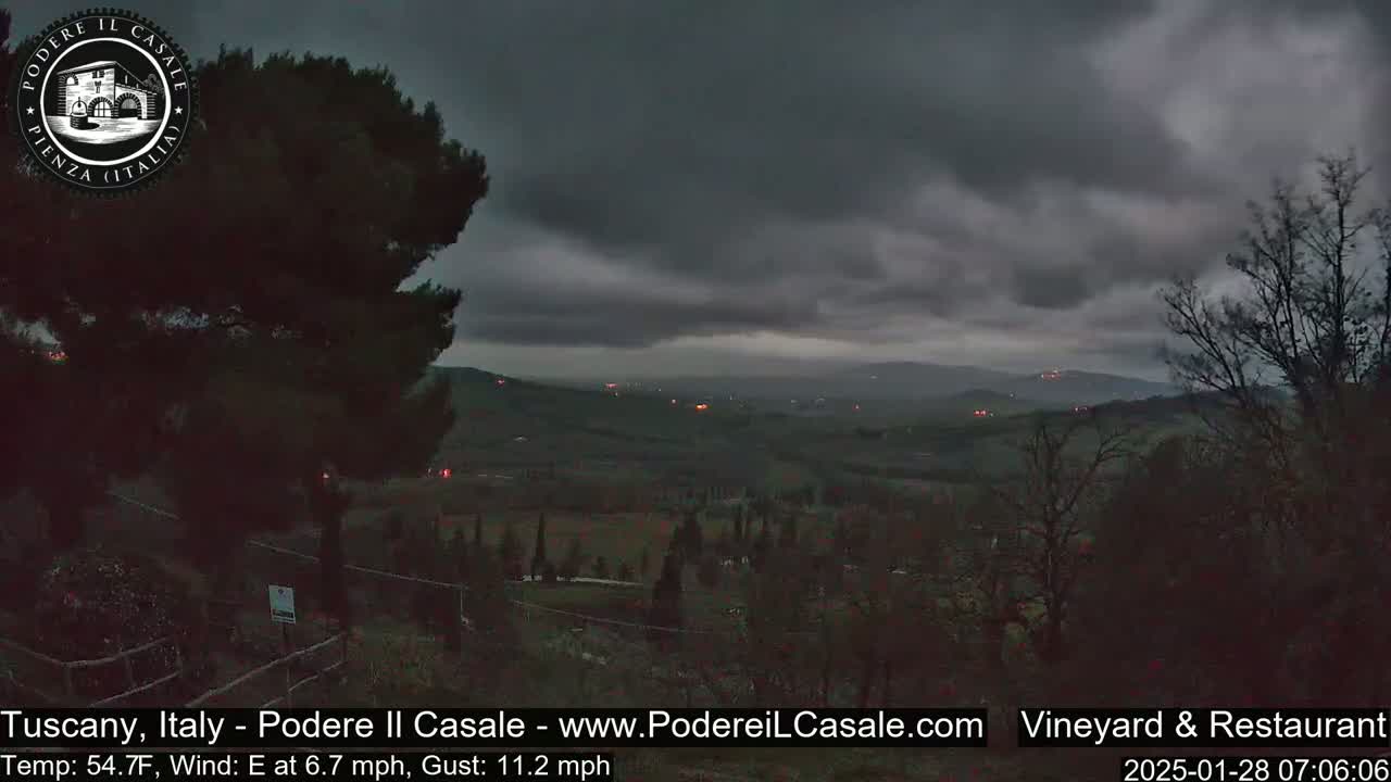 A dark, overcast sky hangs over a hilly landscape dotted with dark trees and a few distant lights.